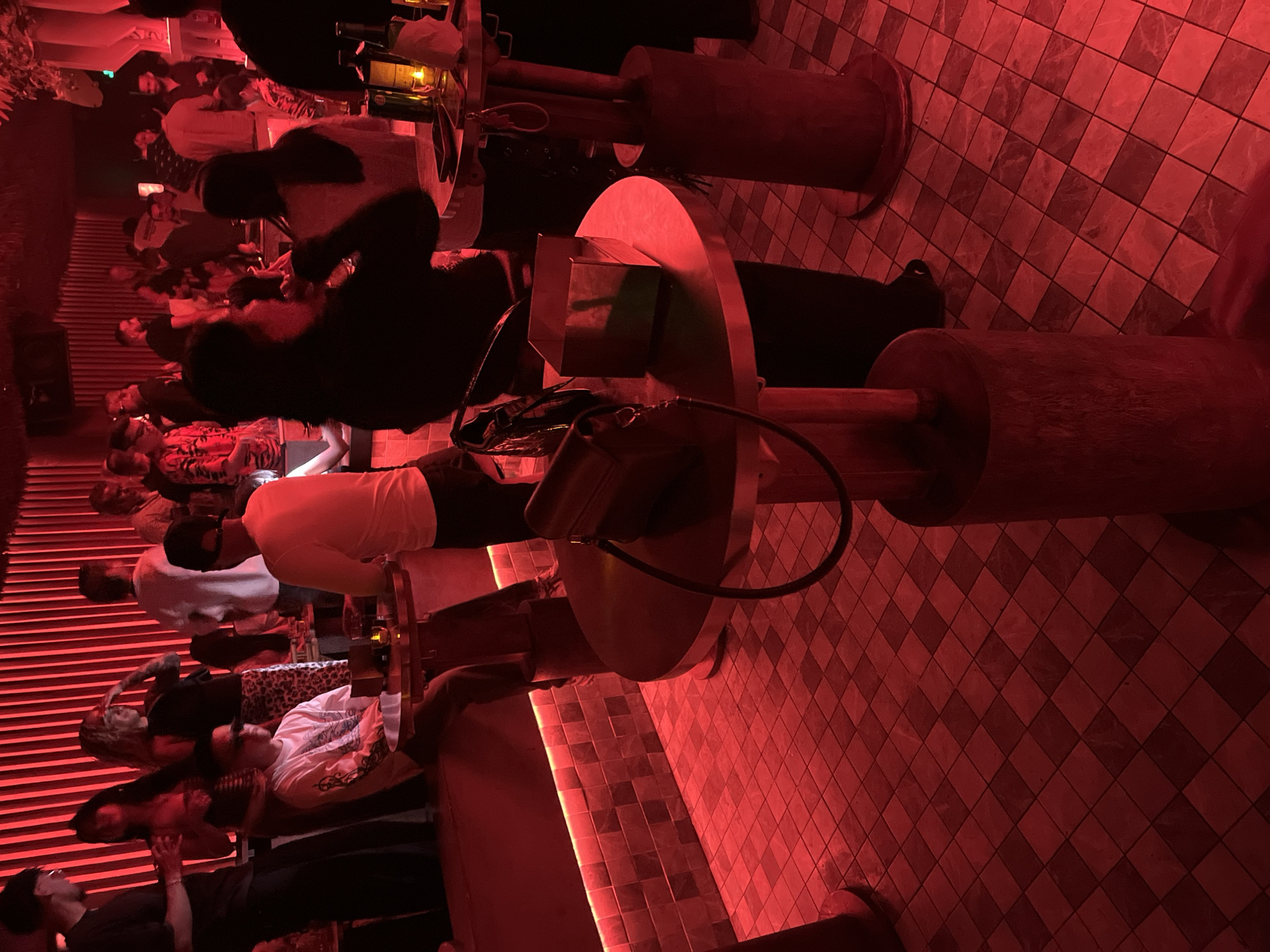 The image depicts a crowded indoor setting with red lighting and diamond-patterned flooring. Several individuals are visible, some seated at cylindrical tables with attached circular platforms. The scene gives the impression of a bar or lounge in Thủ Đức, Vietnam. Objects present include tables, chairs, bags, and clothing worn by patrons.