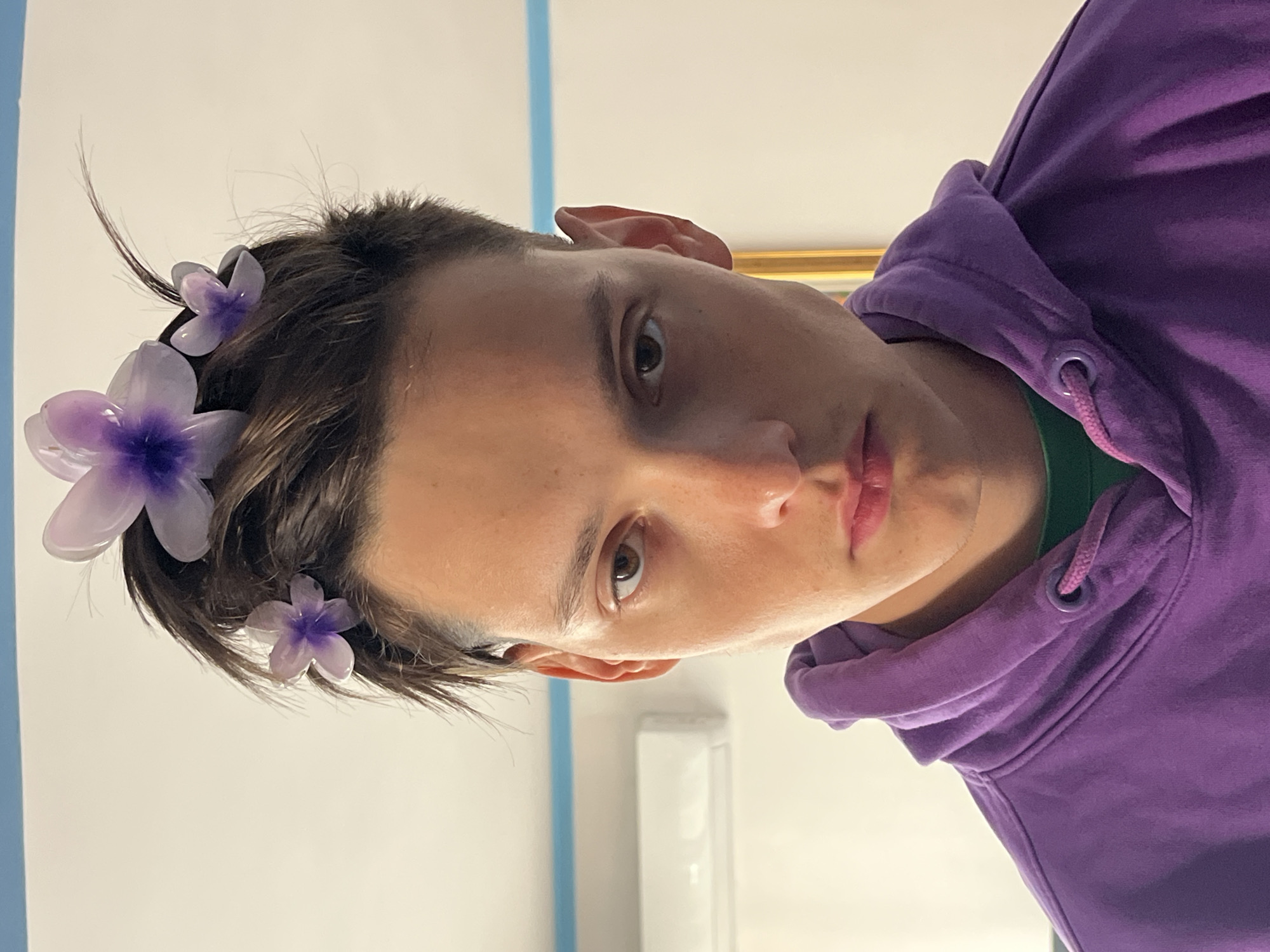 A young person, likely in their late teens or early twenties, is wearing a purple hoodie and has several artificial purple flowers in their hair. They appear to be indoors, in a room with white walls and a blue stripe along the upper portion of the wall. The background suggests a modern interior, possibly a hotel room or apartment. While the location is specified as Budva, Montenegro, there is no visual indication of this in the photo itself.