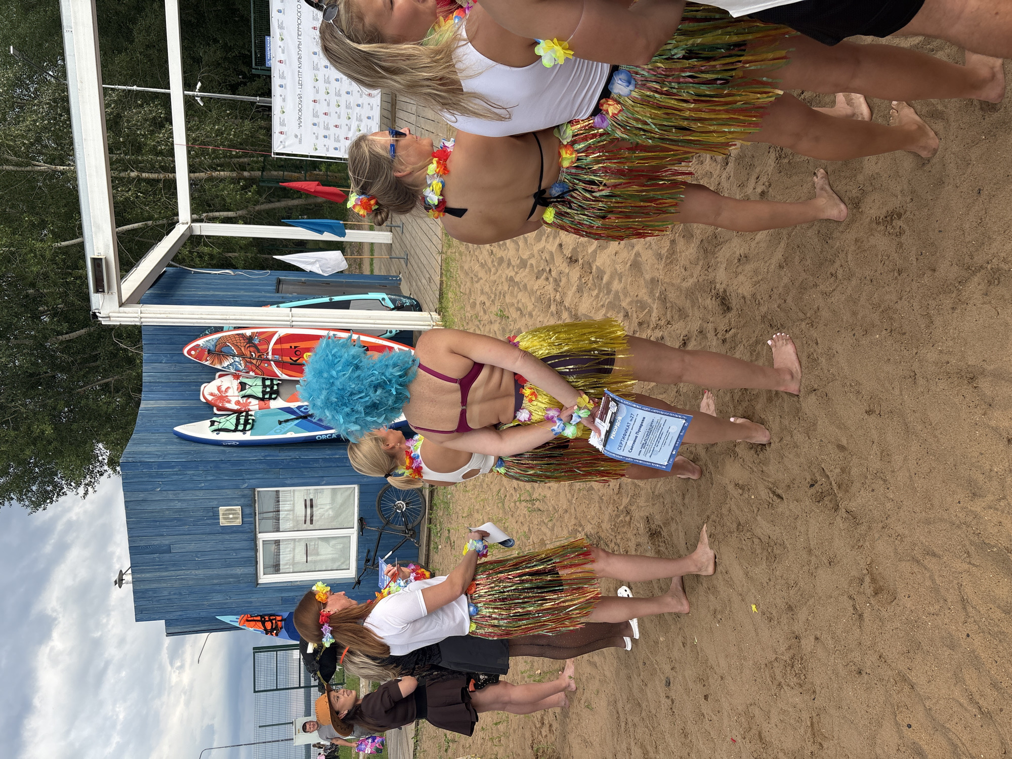 The image shows a group of people, predominantly women, standing on a sandy area, likely a beach, in front of a blue wooden building. Several women are wearing Hawaiian-themed attire, including lei necklaces and grass skirts. Some of the women are holding certificates. Surfboards are visible leaning against the building. In the background, trees and a cloudy sky are present. A sign on the right side of the image contains Russian text, which, based on context clues such as the surfboards and beach setting, might indicate a water sports or recreational facility. The location is identified as the city of Chaikovsky, Russia, likely at a waterfront recreational area.