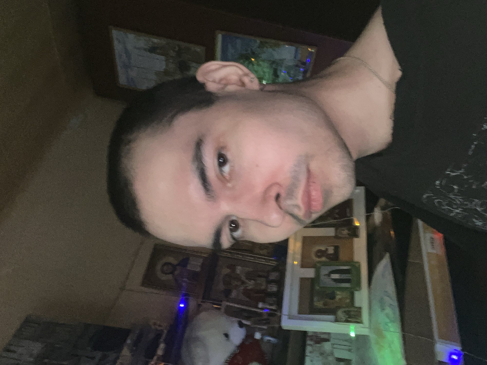 The image is a selfie of a person with short dark hair and light skin. They are wearing a black t-shirt and a silver chain necklace. The background appears to be a room with several framed religious icons and some decorative lights, suggesting a personal or religious space. There is no information in the image that would allow for the determination of the location being Kazan, Russia. The scene is indoors, and the lighting is dim. The focus of the image is the individual's face and upper torso.