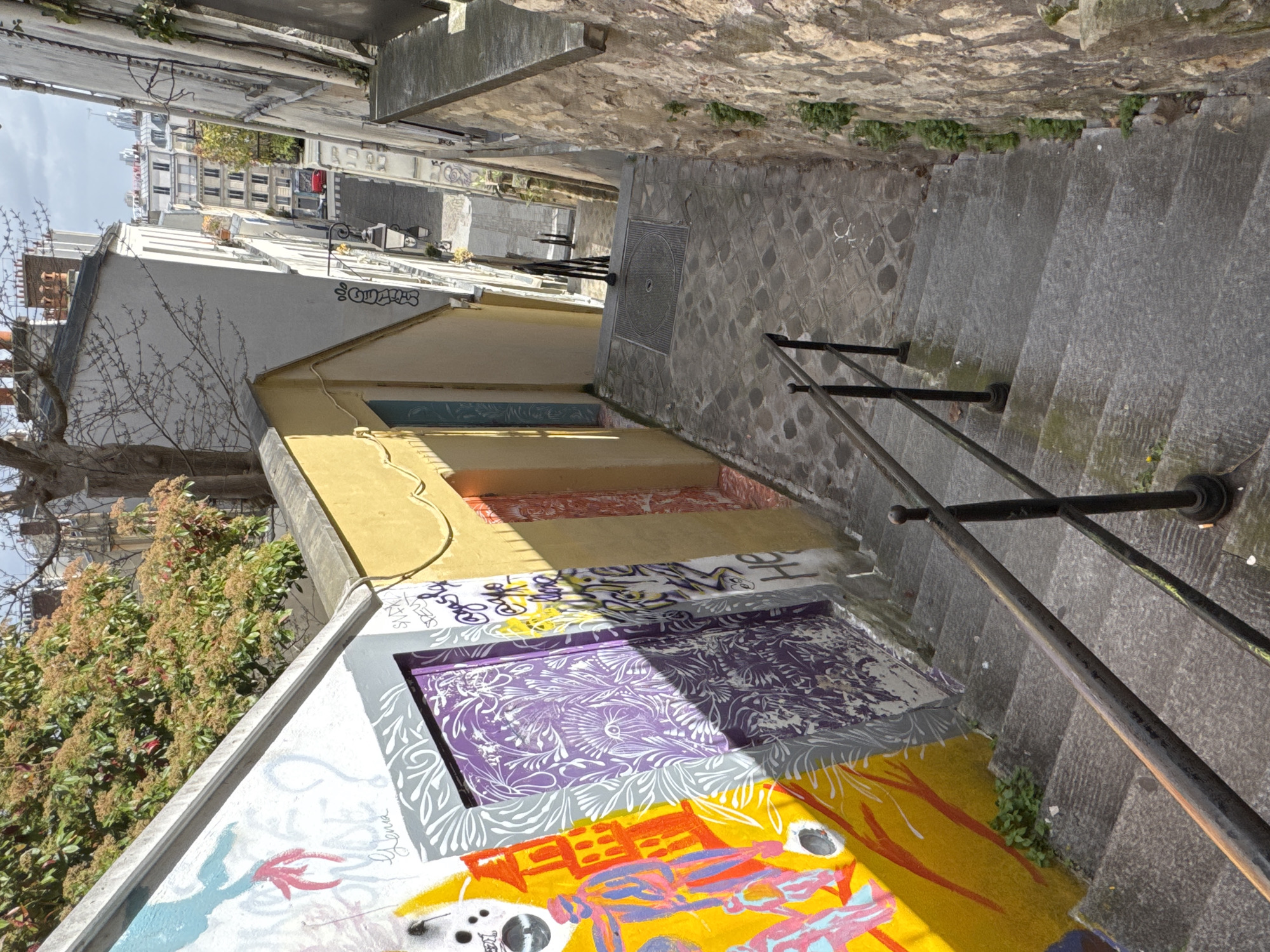 This is an exterior shot of a stairway in the city of Paris, France. The perspective is looking down the stairs. A black metal handrail runs along the right side of the stairs. The walls adjacent to the stairs are covered in colorful graffiti art, featuring a variety of patterns and images. Buildings line the street at the top of the stairs. The street is paved with stone. The sky is overcast.