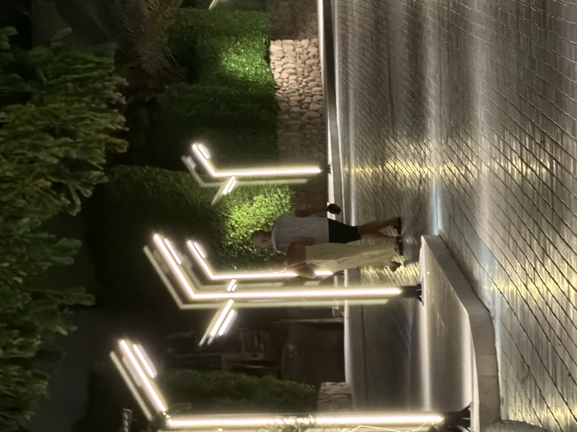 A couple walks along a pathway illuminated by modern, geometric streetlights at night in Sharm El Sheikh, Egypt. The pathway is paved with textured stones, reflecting the bright lights. Lush green bushes and a stone wall form the background, with the branches of trees overhead creating a canopy. The man is wearing a white t-shirt and dark shorts, while the woman is in a flowing, light-colored dress. They are walking away from the camera, side by side.