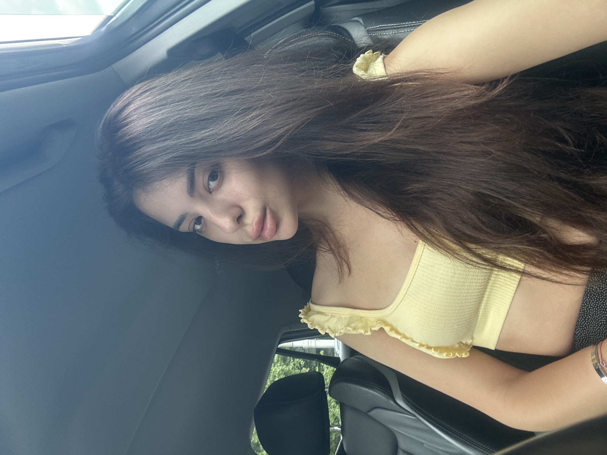 This media file features a young woman posing for a selfie inside a car. She has long, dark brown hair, parted slightly to one side, flowing around her head. Her expression is neutral to slightly pouting, and she looks directly at the camera. She is wearing a light yellow, ribbed or smocked crop top with ruffled straps. On her left wrist, a silver-toned bracelet with some faint, illegible markings is visible. Her lower garment appears to be a dark, possibly black, fabric.
The setting is the interior of a car, with dark, possibly leather, seats visible. Bright natural light streams in from above, indicating it is daytime. A partial view through the car window reveals green foliage, suggesting trees, and a light-colored sky, implying clear or slightly overcast weather outdoors. The image is taken at an unconventional angle, giving a slightly tilted perspective, likely from a handheld device. The scene takes place in Plovdiv, Bulgaria.