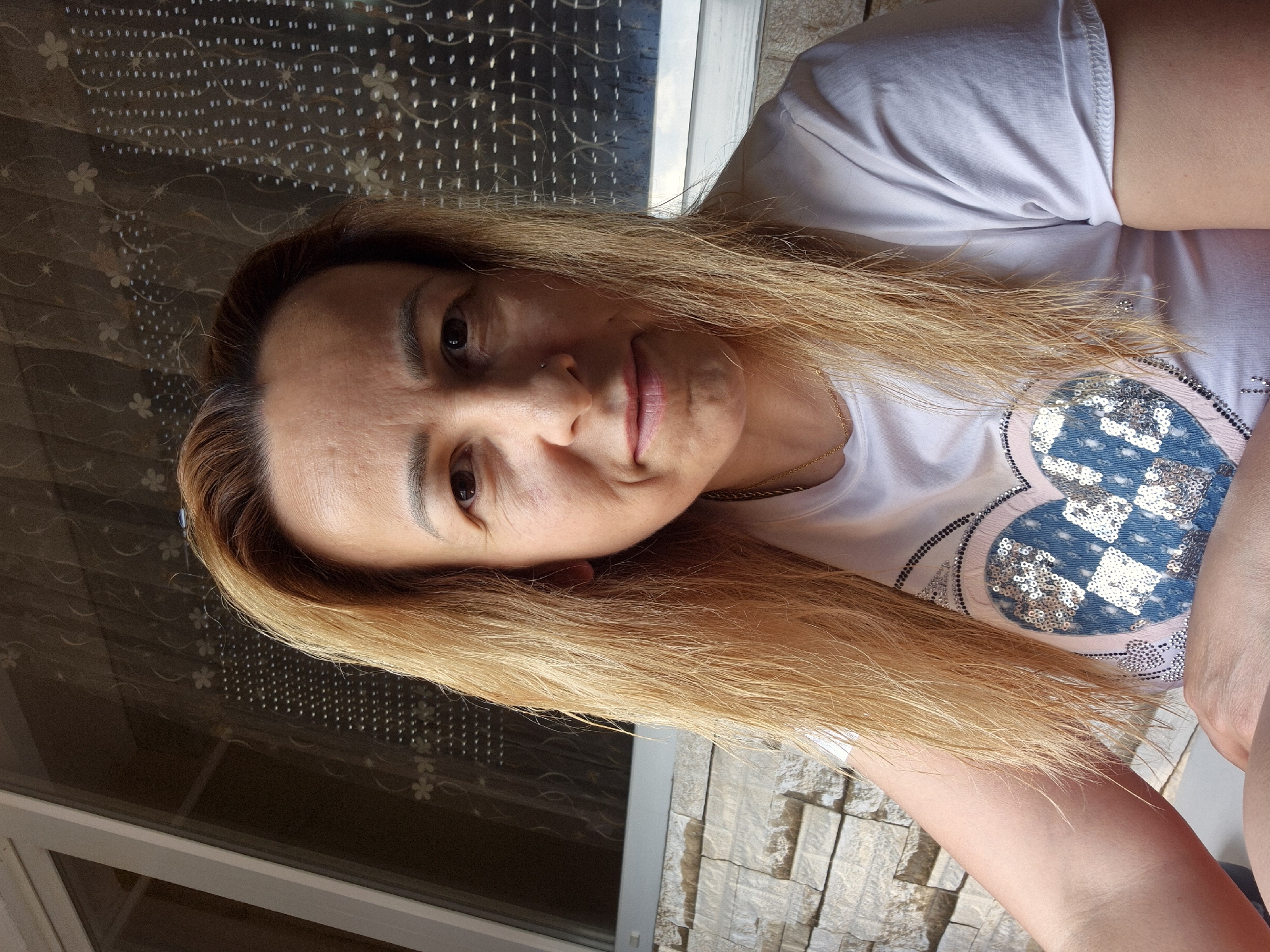 A medium shot shows a woman from the chest up, with her face angled slightly to the right. She has blonde, straight hair that reaches her shoulders and a white t-shirt with a sequined heart design. The background includes a stone wall and a window with a patterned curtain. The lighting suggests an outdoor setting during the day.