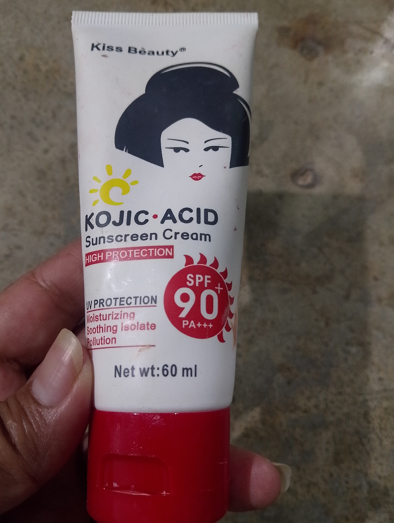 A white tube of Kiss Beauty Kojic Acid Sunscreen Cream with a red cap is held in a person's left hand. The tube features a stylized illustration of a woman's face with black hair and red lips. Text on the tube indicates "HIGH PROTECTION", "SPF 90 PA+++", "UV PROTECTION", "Moisturizing", "Soothing Isolate", "Pollution", and "Net wt: 60 ml". The background appears to be a textured, brownish surface. The image was captured at an angle, showing the front of the tube and part of the person's fingers and nails. The location is the city of Serbelawan, Indonesia.