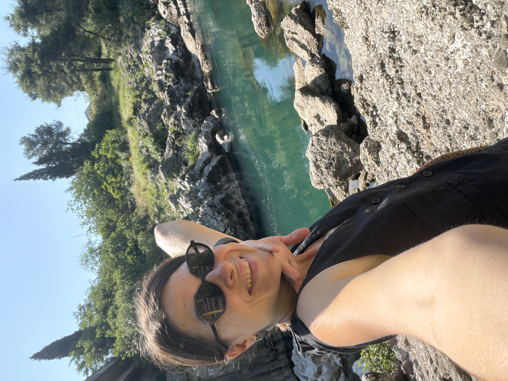 A woman is taking a selfie outdoors by a river. She is smiling and wearing sunglasses and a black sleeveless top. The river is turquoise and surrounded by rocks and trees. A bridge is visible in the background. The image appears to be taken on a sunny day. The specific location is the Skadar Lake National Park, near Podgorica, Montenegro.