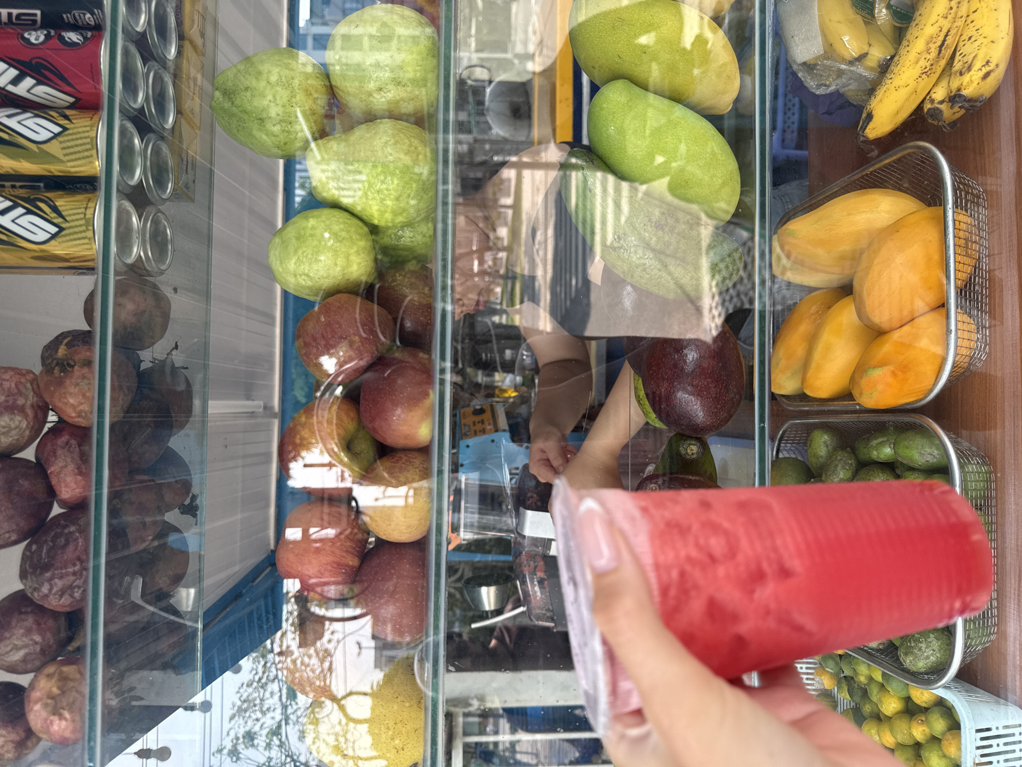 The photograph shows a glass display case filled with various fruits and beverages, including guavas, apples, passion fruits, bananas, mangos, avocados, and canned drinks. A hand is holding a red beverage in front of the case. The location appears to be a street vendor or small shop in Đà Nẵng, Vietnam.