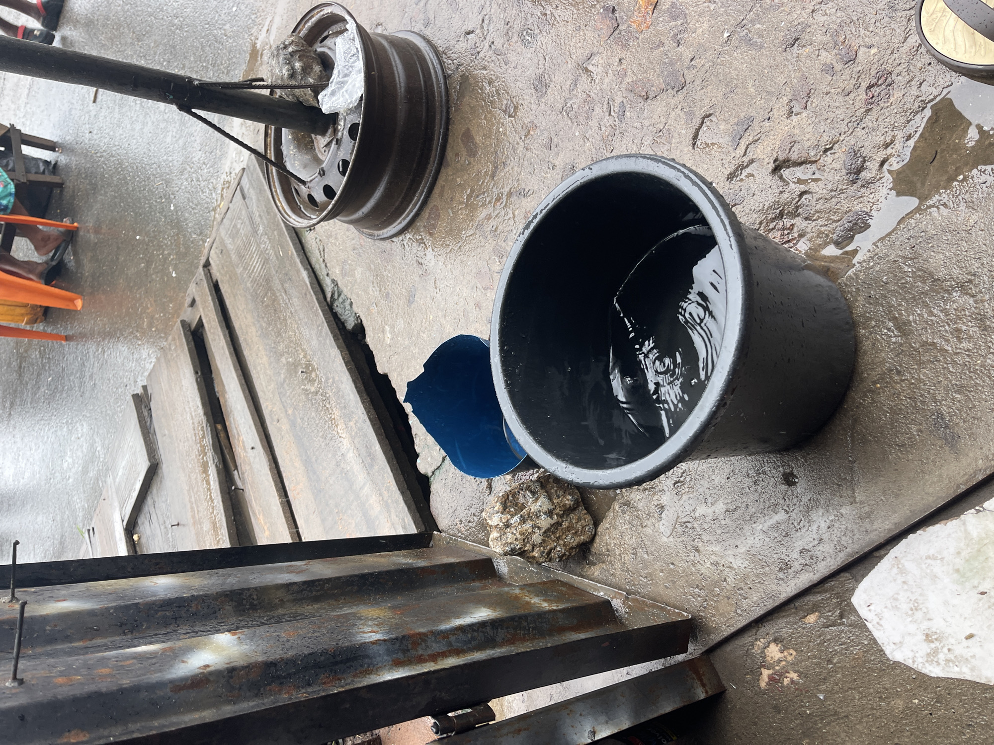 A medium shot captures a street scene in Fegge, Nigeria, on a rainy day. In the foreground, a black plastic bucket is filled with water, with ripples suggesting recent disturbance. To the left of the bucket, a large, rough-textured rock sits on the wet concrete ground. Behind the rock and bucket, a broken blue plastic funnel is visible. Further back, a metal structure, possibly part of a stall or gate, stands on the left side of the frame, showing signs of rust and wear. A pathway made of wooden planks extends into the background, also wet from the rain. In the upper right of the image, a car wheel rim serves as a base for a metal pole, which is secured with rope. In the background, blurry figures are seated, with orange chairs suggesting a seating area. The overall environment is wet and appears to be an outdoor market or commercial area.