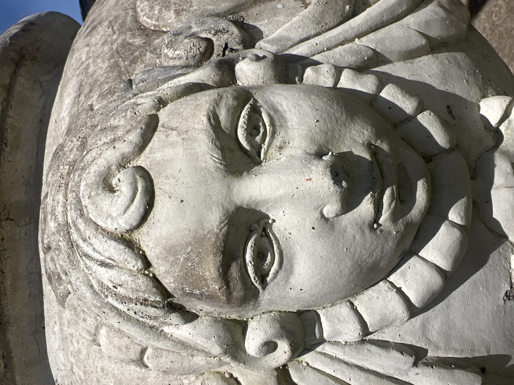 This is a close-up, eye-level shot of a weathered, light-colored stone carving of a child's face on the side of a large urn or pot. The carving features curly hair framing the face, prominent eyes with detailed irises, a small nose, and slightly parted lips. The texture of the stone is rough and shows signs of age and exposure to the elements, with some dirt and discoloration. The background is a blur of light and shadow, with hints of blue sky visible at the top right. The carving appears to be an architectural or decorative element, possibly located outdoors in a garden or public space in Pa Khlok, Thailand.