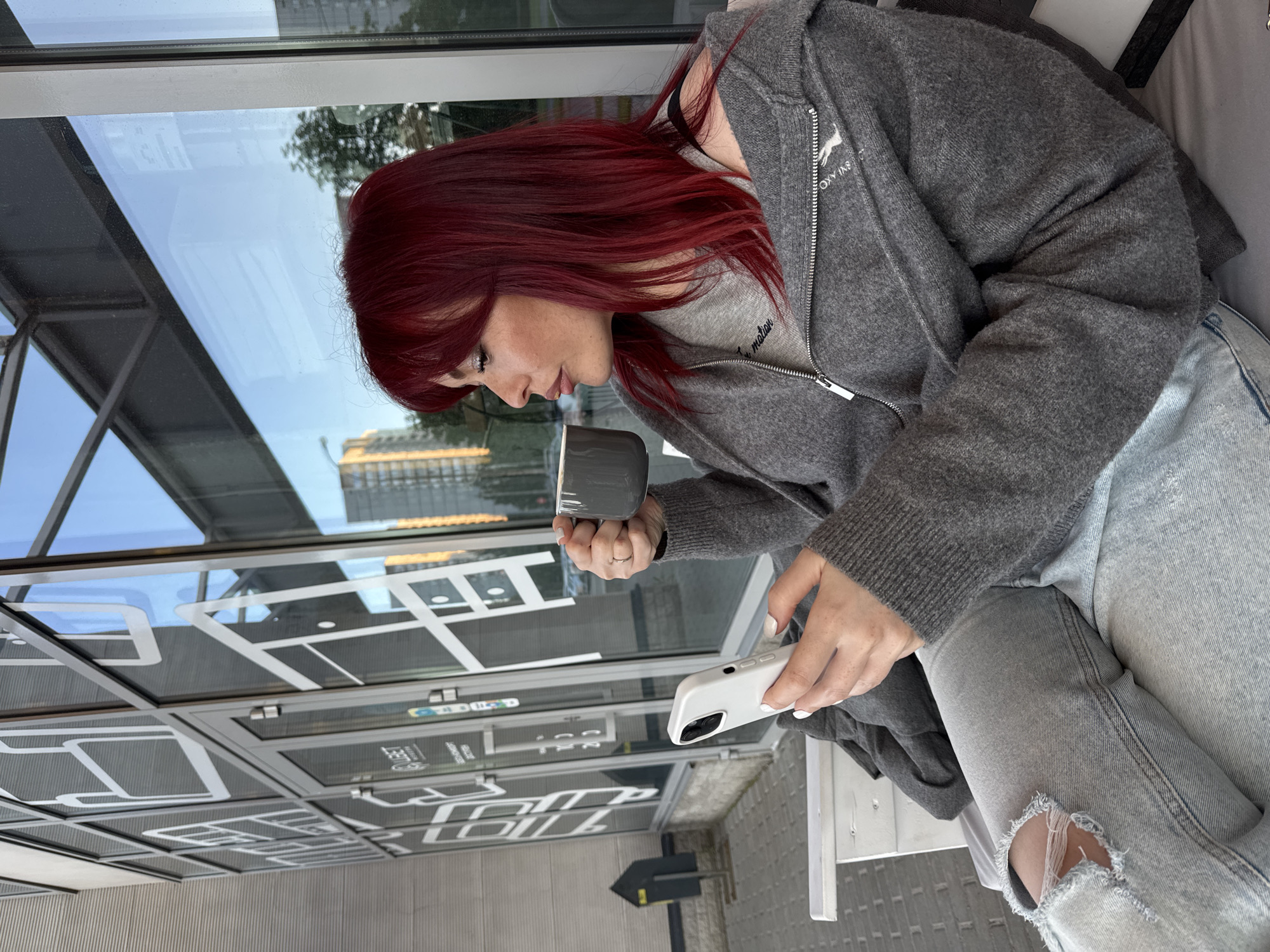 A young person with long, wavy red hair is captured in a profile shot, looking down intently. They are seated, dressed casually in a grey hooded zip-up sweater with a lighter grey shirt underneath, and light-wash ripped jeans. In their right hand, they hold a grey mug, poised as if taking a sip, while their left hand holds a white smartphone, with white nail polish visible on their fingers. A simple ring is worn on the left ring finger.
The setting appears to be outdoors or in a covered area, possibly a cafe or public space, bordered by large glass panels. These panels feature white, linear graphic designs that resemble a stylized map or abstract pattern. Reflections in the glass show glimpses of a modern building with multiple windows and some greenery, indicating an urban or commercial environment. The light suggests it is daytime with clear weather.
Visible text on the grey sweater includes a white logo that partially reads "ROXY INS" or similar, with a small animal design (possibly a fox or deer). Below this, on the lighter grey inner shirt, partial text is visible, possibly "motion". Although not clearly legible as specific words, the white linear patterns on the glass panels contribute to the modern aesthetic of the surroundings. The person appears relaxed and focused on their drink and phone.