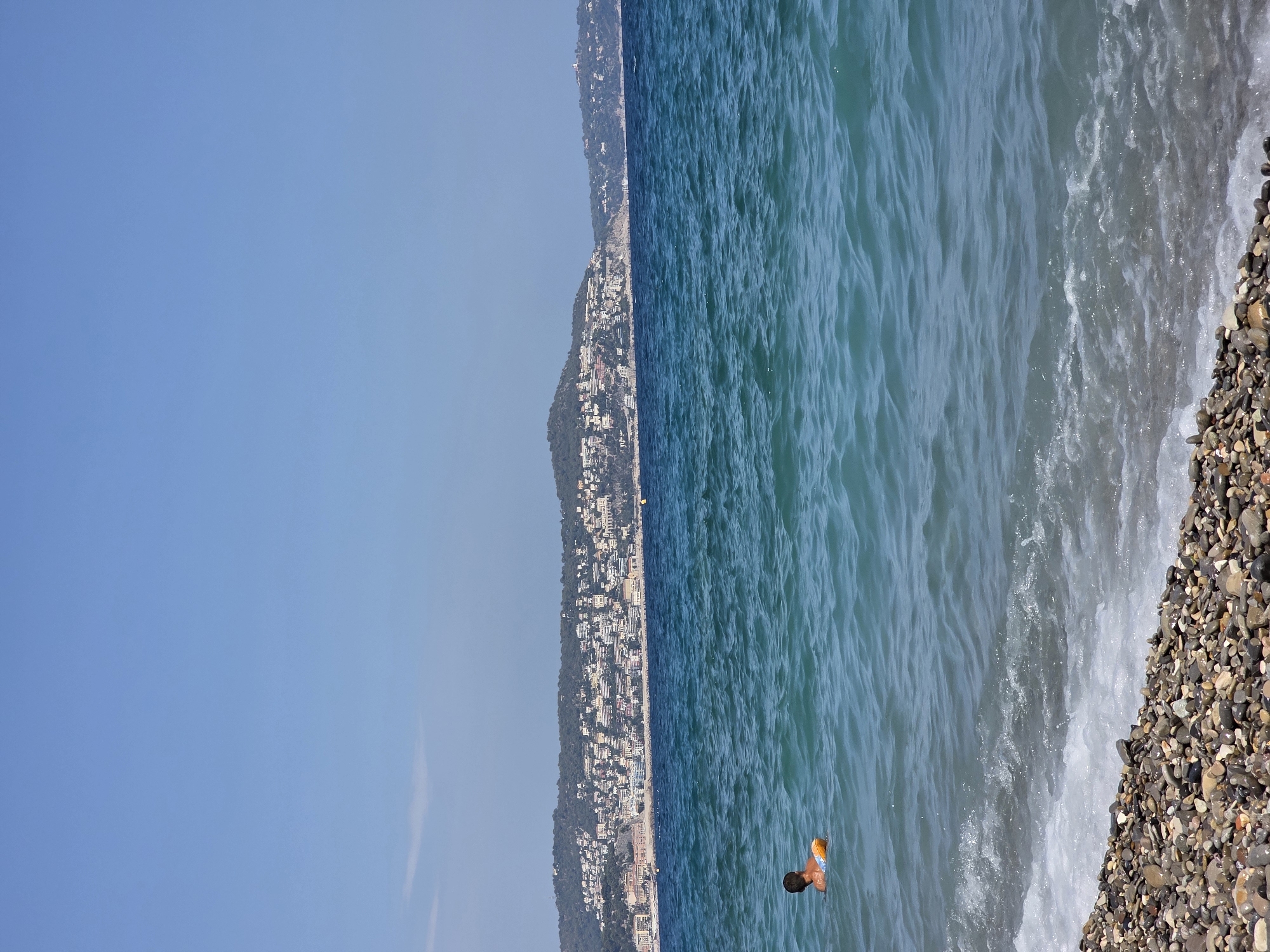 This is a vertical photograph of a coastline with a city built on a hill overlooking a body of water. The sky is clear and blue, suggesting a sunny day. The water is a deep blue-green and appears to have moderate waves, with white foam near the shore. The shore itself is a pebble beach, with various shades of grey, brown, and beige stones.
In the water, a person is swimming, with their head and shoulders visible above the surface. They are wearing swim trunks that appear to be orange and blue. The person is positioned in the right half of the frame, about a third of the way down from the top.
The coastline is lined with a densely packed city, characterized by numerous buildings in white and pastel colors. The city extends up the hillside, which is covered with lush green trees. The overall impression is a vibrant coastal resort town on a clear, bright day. The scene is peaceful and suggests a recreational activity like swimming or enjoying the beach. There is no visible text in the image.