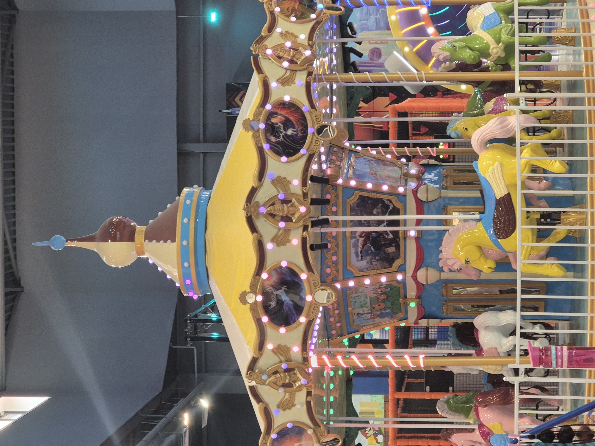 A brightly lit indoor amusement center in Kokshetau, Kazakhstan, features a carousel. The carousel’s structure is adorned with yellow and white decorative panels, and it has painted horses and other animals. The scene is viewed from a low angle, emphasizing the height of the structure. White bars surround the carousel. Multi-colored play structures are visible in the background. The ceiling of the building is visible above, and has lights.