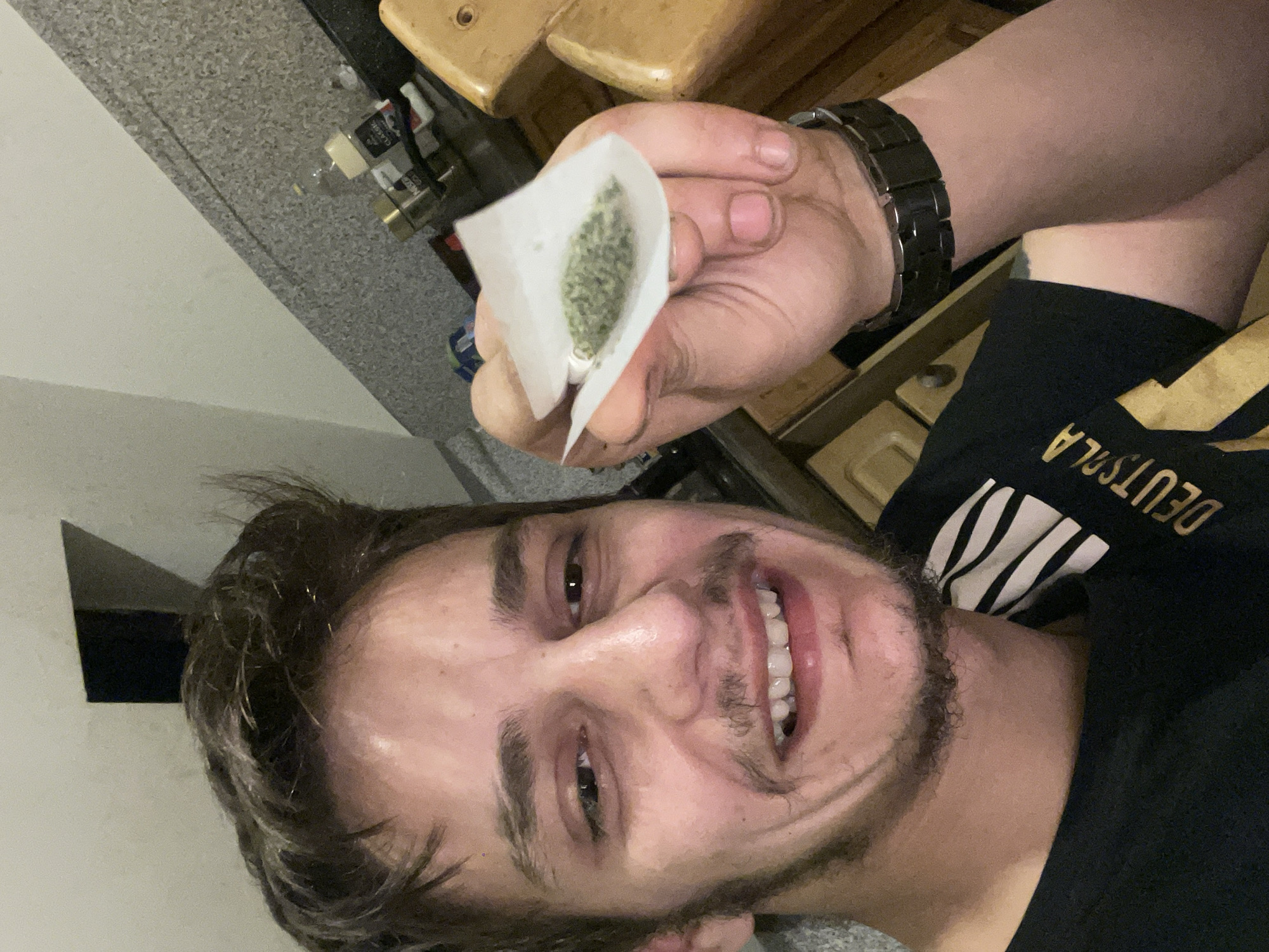 A man is holding a folded piece of paper filled with what appears to be marijuana. The man is light-skinned with dark hair and a beard, wearing a black t-shirt with gold lettering and a black watch. The background includes a gray wall, wooden furniture, and what appear to be household items. The location is unknown.
