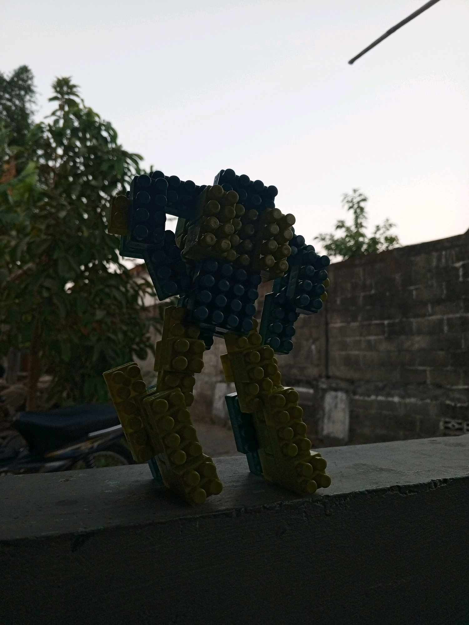 A toy robot constructed from yellow and blue plastic building blocks stands on a grey concrete ledge in Bantul, Indonesia. The robot is positioned in profile, facing left, with its legs bent and its arms raised slightly. Behind the robot, a brick wall runs horizontally across the frame. To the left of the wall and robot, a leafy green tree is visible, and further to the left, the dark silhouette of a motorcycle is present. The sky above is a pale, overcast grey. The overall lighting suggests either dawn or dusk.