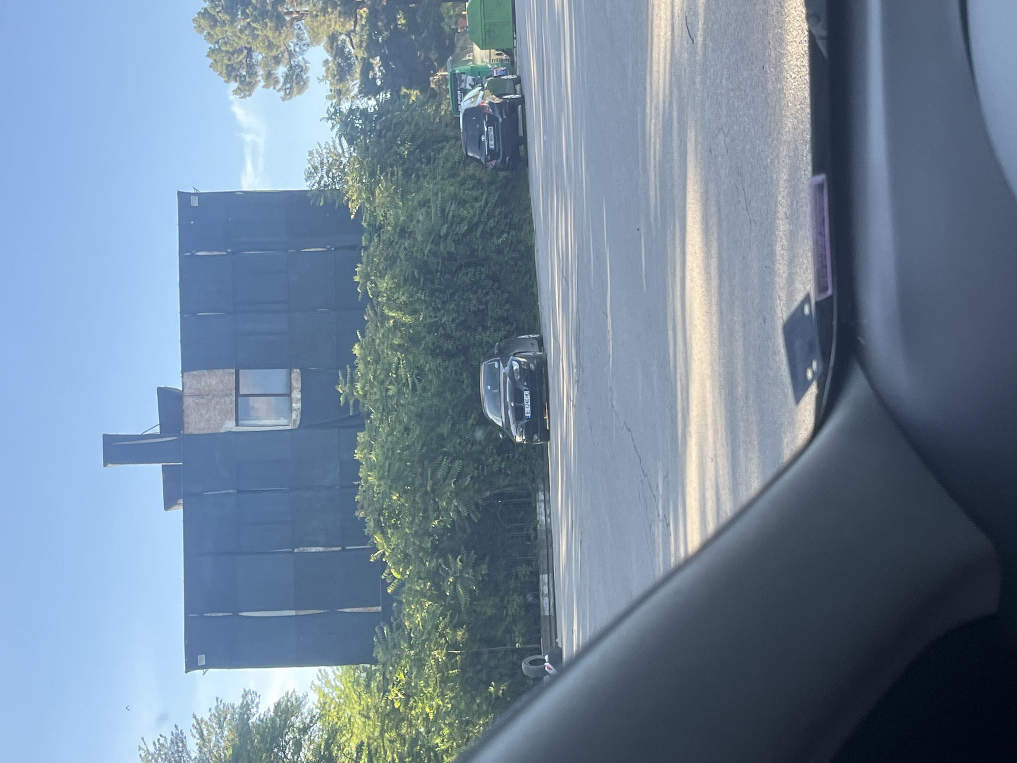 The image shows a view from inside a vehicle, likely a car, looking towards a building under construction or renovation. The building is tall, with a dark protective covering over most of its facade, except for a section that appears to be unfinished or temporarily exposed. Next to the building, there are numerous trees and foliage. Parked along the road adjacent to the trees, there are at least two vehicles. The image appears to have been taken on a sunny day, indicated by the bright sky and shadows cast on the road. The location is Blagoevgrad, Bulgaria.