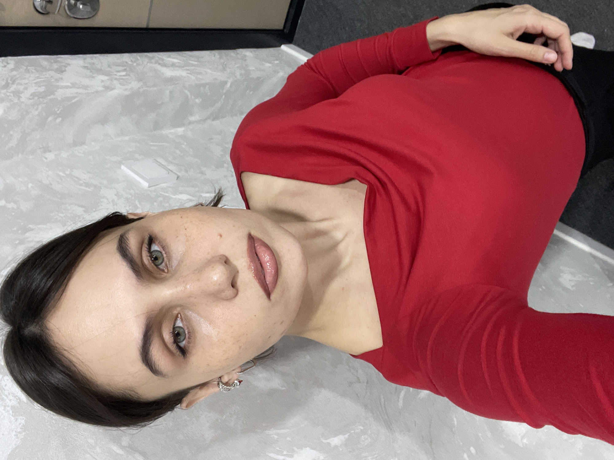 A young woman with fair skin and dark hair is lying on a light gray surface, possibly a floor, wearing a red long-sleeved shirt. Her eyes are a light green color, and she is wearing lip gloss. She has a small earring in her left ear. Her right hand is visible near her waist. The background includes a dark object, potentially a piece of furniture. The location is not evident from the image.