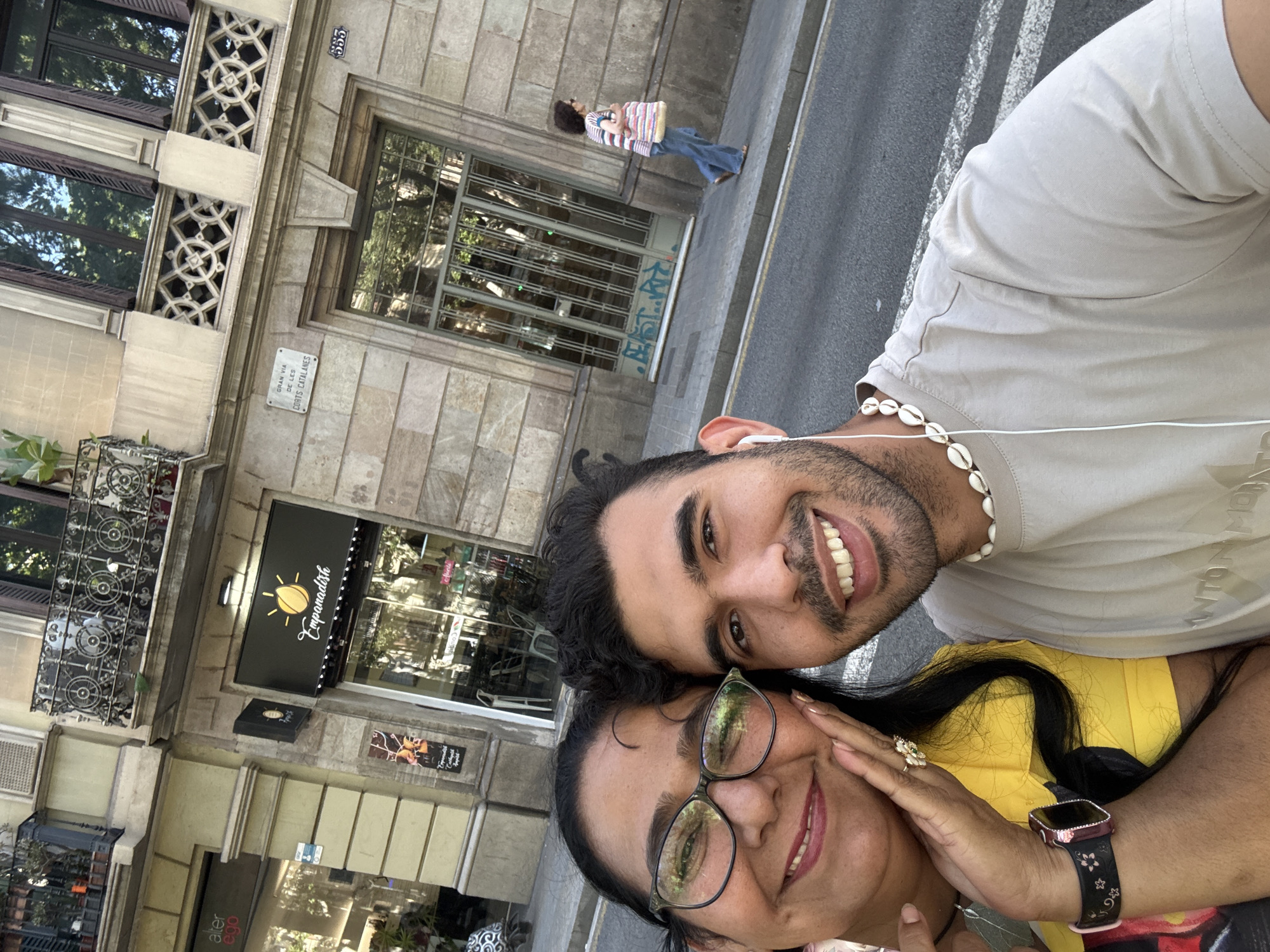 This is a selfie taken outdoors in the city of Barcelona, Spain, on a street with a building facade and a sidewalk. In the foreground, two people, a woman and a man, are posing for the photo. The woman, on the left, has dark hair, wears glasses, and is smiling. She has her right hand on her cheek and is wearing a yellow shirt and a watch. The man, on the right, has dark hair and a beard, is wearing a light-colored t-shirt and a shell necklace, and also smiling. Both are looking towards the camera.
Behind them, the building facade is made of stone blocks, with ornate balconies and large windows. A sign on the building reads "Empanadish". A street sign indicates "GRAN VIA DE LES CORTS CATALANES". A woman with dark curly hair, wearing a striped shirt and jeans, is walking on the sidewalk in the background, carrying a bag. The street in front of the building is paved and has white lines.