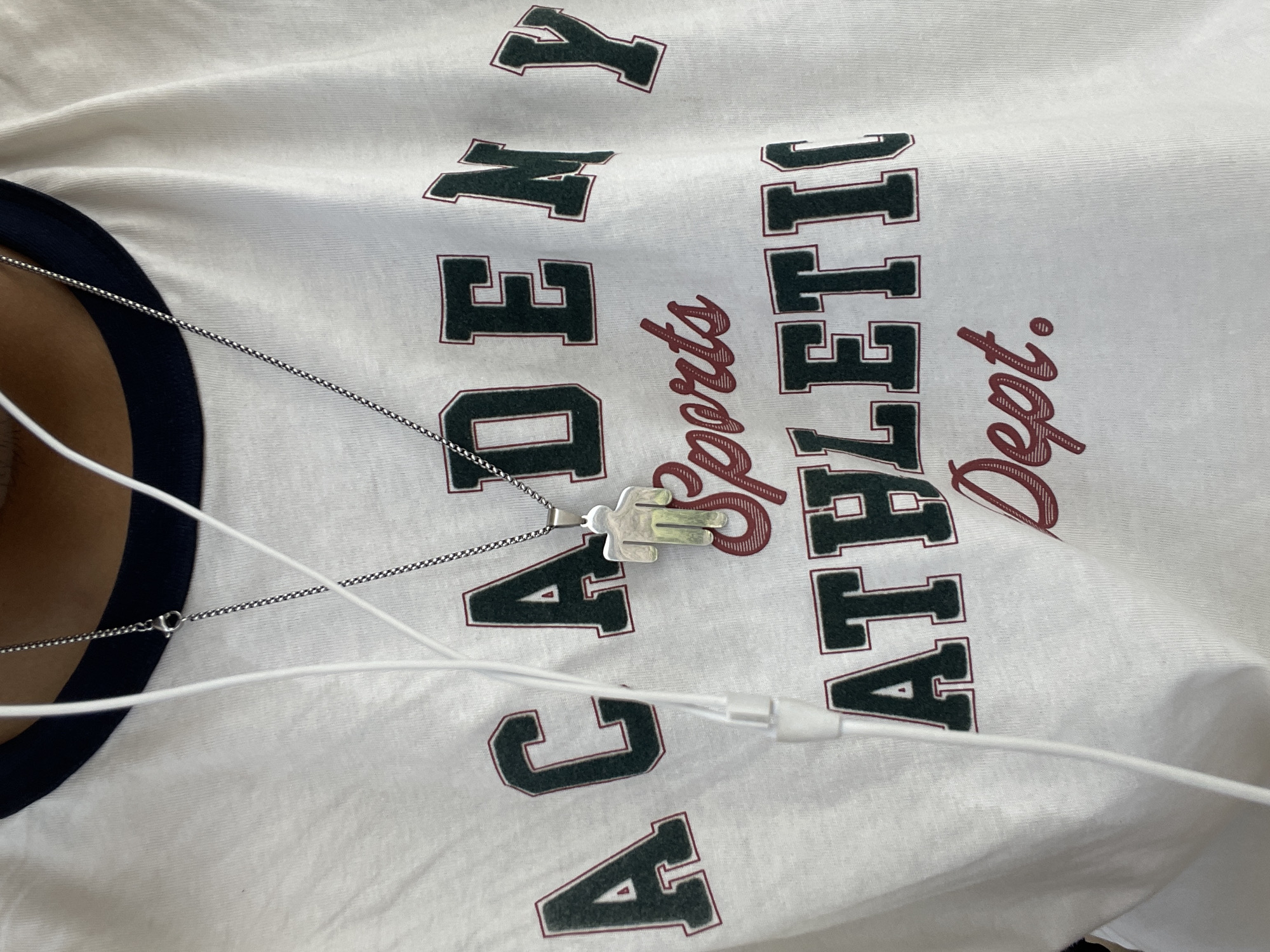 A close-up shot shows a person wearing a white t-shirt with a dark blue neckline. The t-shirt has text printed on it in dark green and maroon. The top line reads "ACADEMY", the middle line says "Sports" in script, and the bottom line reads "ATHLETIC DEPT.". A silver chain necklace with a small, abstract human-shaped pendant hangs over the t-shirt, resting on the word "ACADEMY". White earbud wires are visible in the foreground, partially obscuring the text on the shirt. The lighting suggests an outdoor setting, possibly during the day. No specific location in El Valle del Espíritu Santo, Venezuela can be identified from the image.