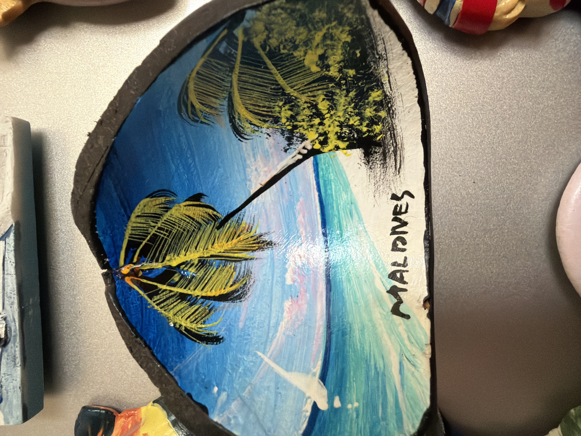 The image shows a close-up of a souvenir magnet depicting a beach scene with palm trees and the word "MALDIVES" painted on it. The magnet is attached to a metallic surface, likely a refrigerator. The scene is a painting on what appears to be a shell or a similar natural object, with vibrant colors of blue for the sky and water, green and yellow for the palm trees, and white for the sand. A sailboat is visible in the distance. The surrounding area of the magnet has a dark, irregular border. Other magnets or small decorative items are partially visible in the background and at the bottom edge of the image, including a figure with a red and white striped hat and clothing. The image is taken from a slightly downward angle. There is no information provided about the location in Pazardzhik, Bulgaria.
