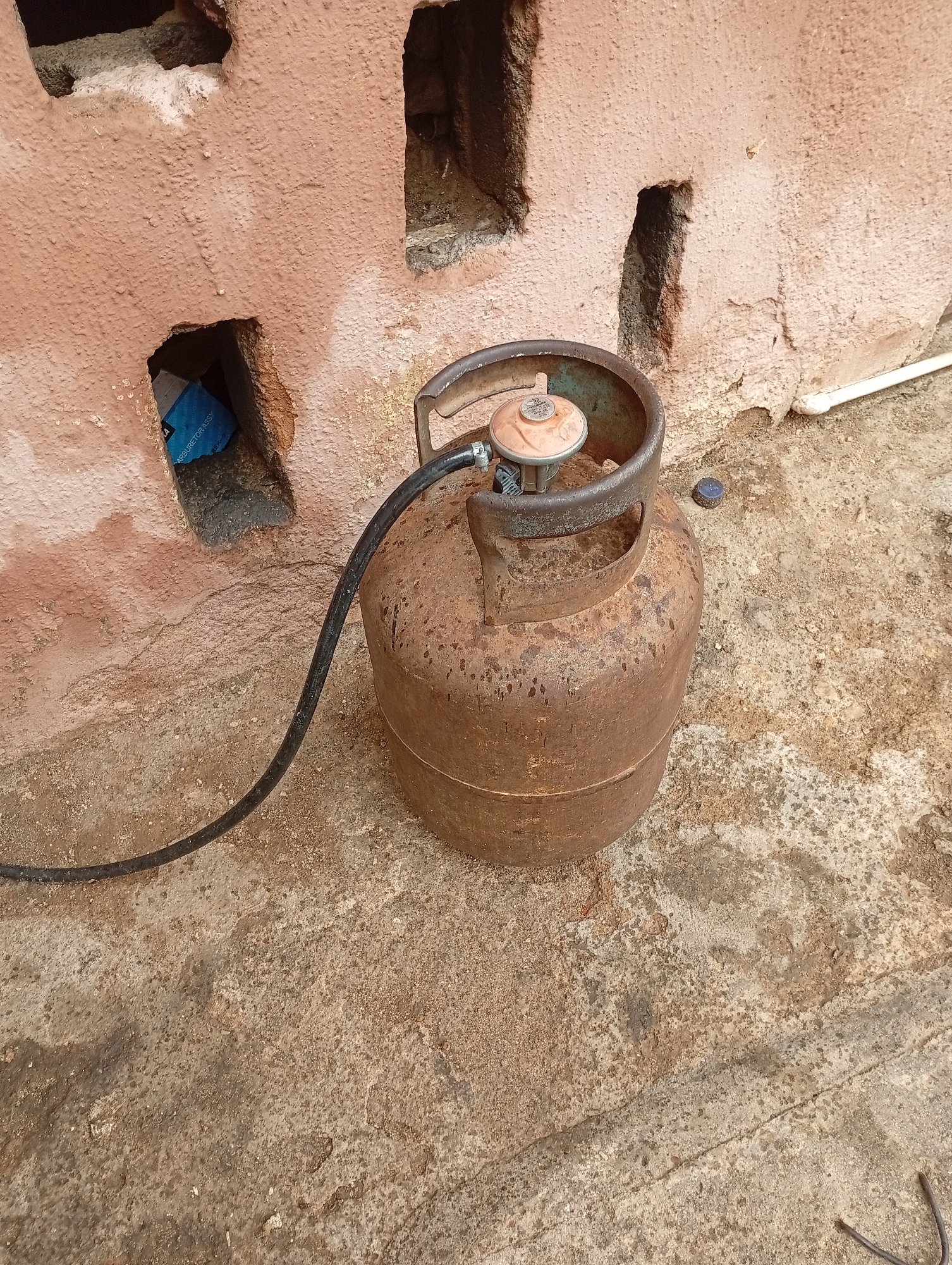 A rusted, old gas cylinder with a black hose attached sits on a worn concrete surface. The cylinder has a regulator attached to the top. Behind the cylinder, a rough, pinkish-colored wall with rectangular openings is visible. In one of the openings, a blue box with white text is partially visible. The environment appears to be outdoors in Rafin Par, Nigeria.