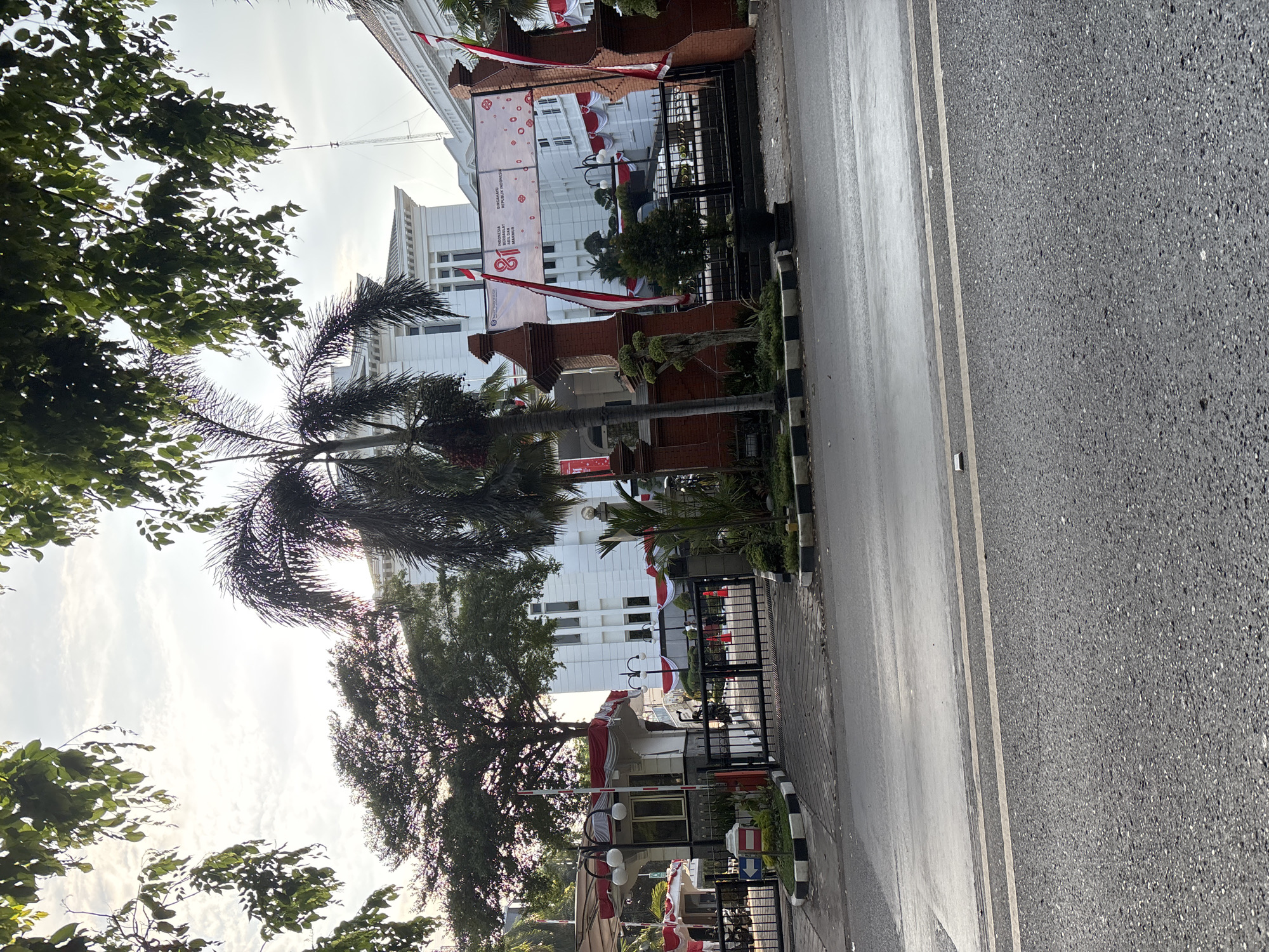 The image depicts the exterior of a white building, likely an administrative or historical structure, in Cirebon, Indonesia. The building features multiple windows and is adorned with red and white banners, suggesting a celebration, possibly Indonesia's Independence Day, given the prominent "81" on one banner. The scene is framed by lush green trees, with a palm tree in the foreground. A black metal gate with brick pillars guards the entrance, and a small security booth is visible to the left. The street in front of the building is wet, indicating recent rain or morning dew. The sky is overcast with visible clouds. The overall impression is one of a well-maintained civic space decorated for a significant national event.