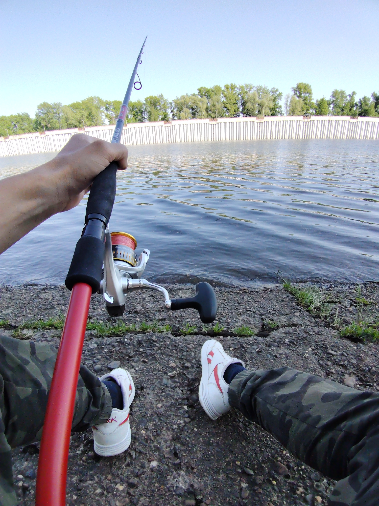 The image shows a first-person view of someone fishing by a body of water. The person is holding a red and black fishing rod with a silver reel in their right hand. The water appears to be a river or lake, with a concrete retaining wall visible on the opposite bank. Trees are visible behind the wall. The person is wearing camouflage pants and white sneakers with red stripes. The ground beneath them is cracked concrete with small patches of grass. The location is likely in the city of Chaikovsky, Russia.
