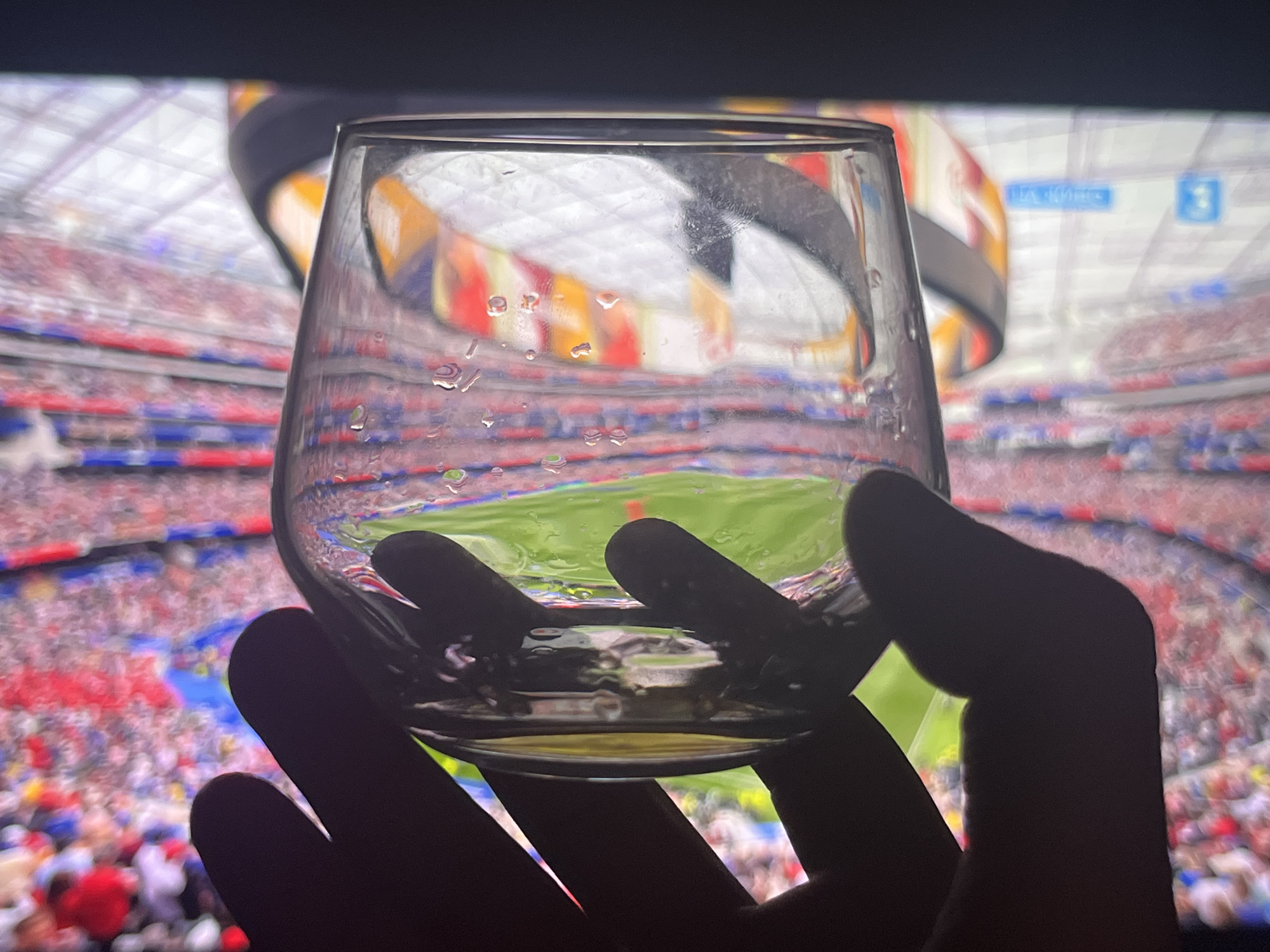 A hand holds a partially filled glass of amber liquid in the foreground, obscuring a television screen displaying a live soccer match. The stadium is packed with a crowd wearing a mix of red, blue, and white attire. The screen shows a vibrant green field with a player in red visible in the background. The glass itself is slightly smudged with condensation droplets. The scene is dimly lit, with the primary light source emanating from the television screen. No specific location in Varna, Bulgaria, can be identified from the image.