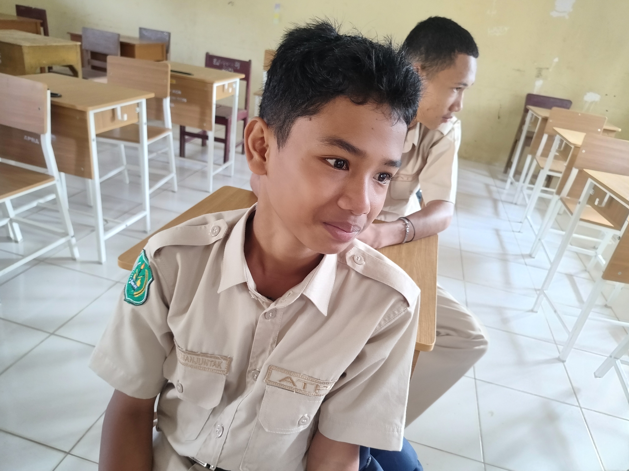 Two young males, likely students, are in a classroom. The student in the foreground, on the left, is wearing a tan uniform shirt with patches and name tags. He has dark, short, spiky hair and is looking off to the right. The student in the background, on the right, is also wearing a similar tan uniform shirt and has short dark hair. He is looking down and to the left. They are seated at wooden desks with white metal frames in a room with a light-colored tiled floor and yellow walls. The classroom appears to be in Lawebekung, Indonesia, as indicated by the presence of desks and student uniforms common in Indonesian educational institutions.