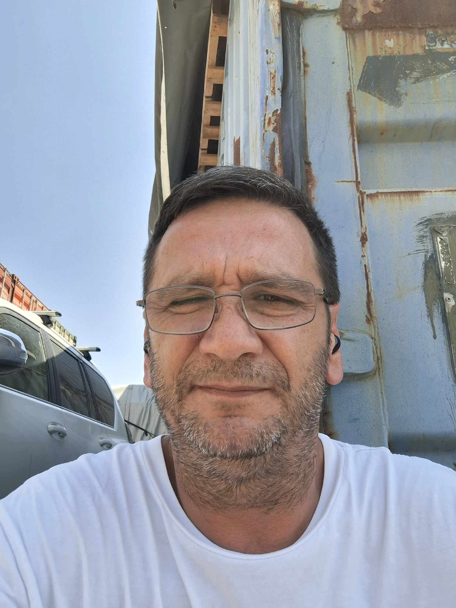 A middle-aged man with short, dark hair, a beard, and glasses stands outdoors in front of a large, rusty, blue shipping container. He is wearing a white t-shirt. The man is looking directly at the camera. To his left, a silver SUV is parked. In the background, there is a clear blue sky and what appears to be more shipping containers and wooden pallets. The scene suggests an industrial or port area. The image is taken in Kiryat Gat, Israel.