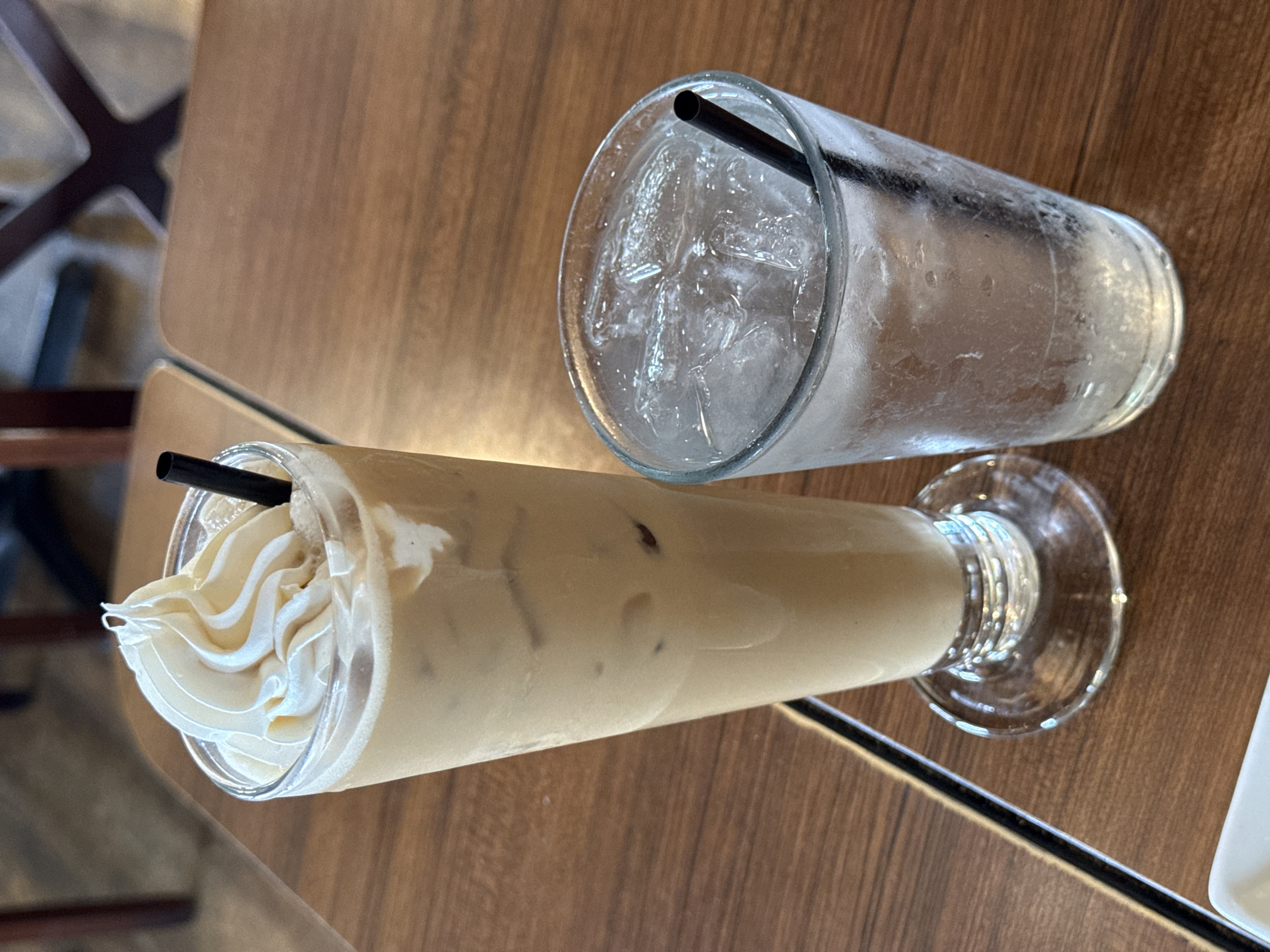 The image shows two glasses on a wooden table. One glass contains a beige-colored beverage with ice and a swirl of white cream on top, along with a black straw. The other glass is filled with ice and water and also has a black straw. The scene appears to be indoors, possibly in a cafe or restaurant. The location can not be determined as San Diego, United States of America, without any additional visual details.