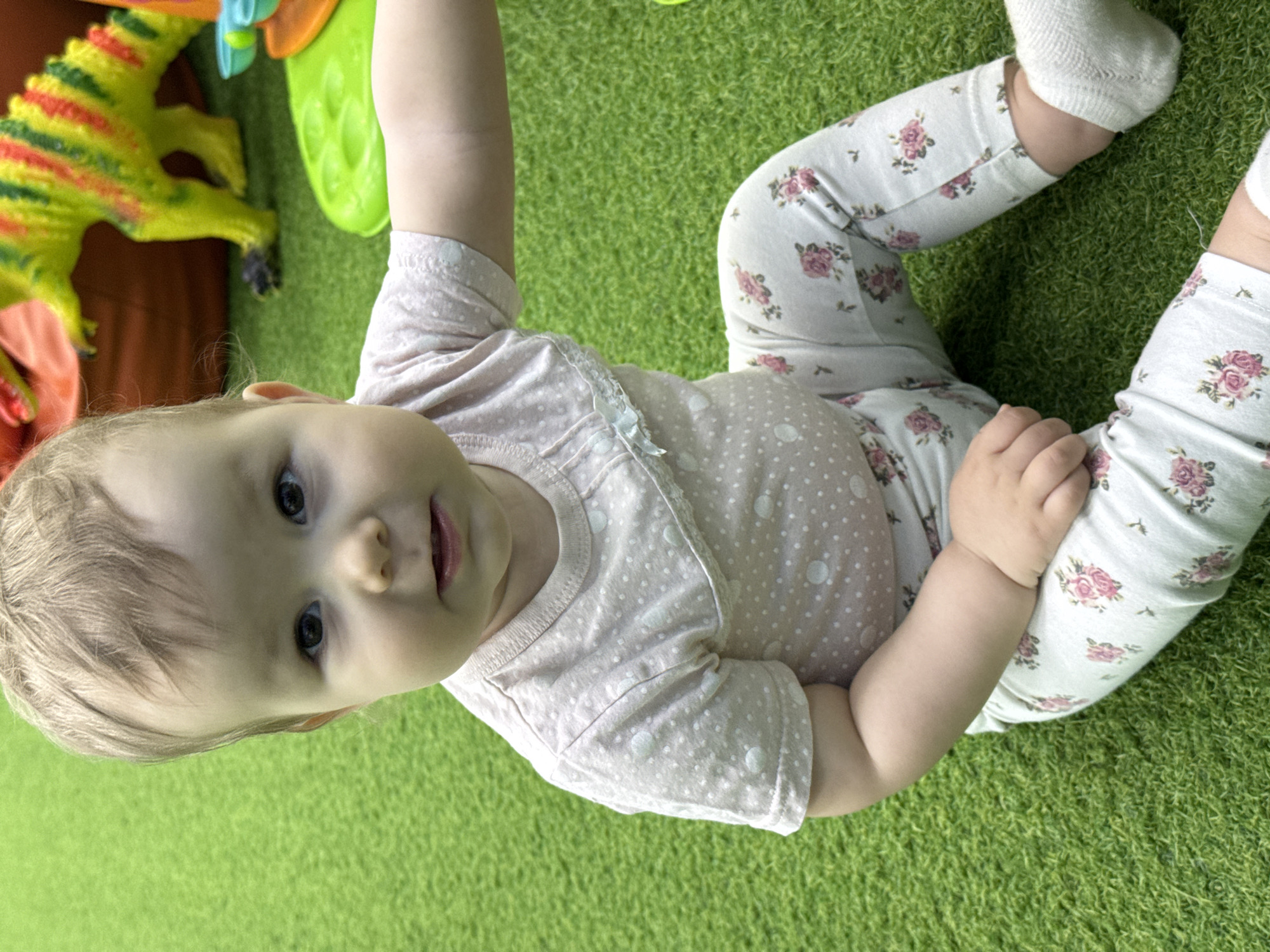 An overhead shot captures a baby lying on vibrant green artificial grass. The central subject is a fair-skinned baby with light blonde hair and dark eyes, looking directly at the viewer with a neutral expression. She is dressed in a light pink top with white polka dots and light-colored leggings featuring a delicate pink rose pattern. White socks cover her feet.
In the upper left corner of the frame, a bright yellow toy dinosaur with red markings is partially visible, indicating a playful environment. Another brightly colored toy, possibly green and orange, is subtly seen at the top edge. The artificial grass creates a consistent, bright green ground cover, suggesting an indoor play area or a dedicated outdoor child-friendly space. The lighting is bright and even. There is no visible text in the image.