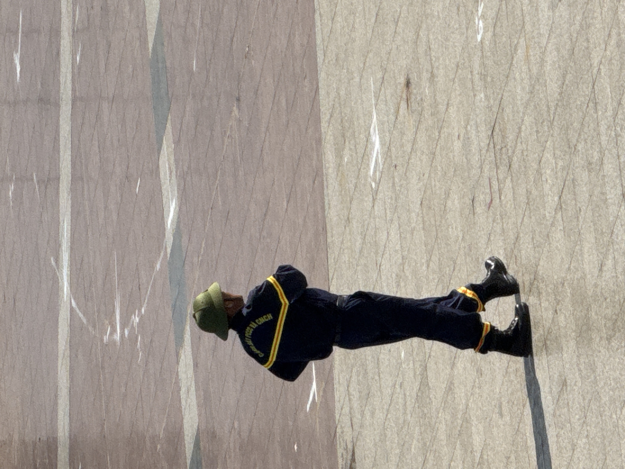 A lone individual in a dark blue uniform with yellow and red trim, and a green helmet, stands on a paved outdoor area. The uniform has text on the back that appears to be Vietnamese. The ground is made of light-colored square tiles. The individual is facing away from the viewer and to the right, with their hands clasped behind their back. The scene suggests an official or guard on duty. The presence of the specific uniform and the paved area is consistent with locations such as the Lam Vien Square in Da Lat, Vietnam, known for its open public spaces and historical significance, often utilized for parades and official events.