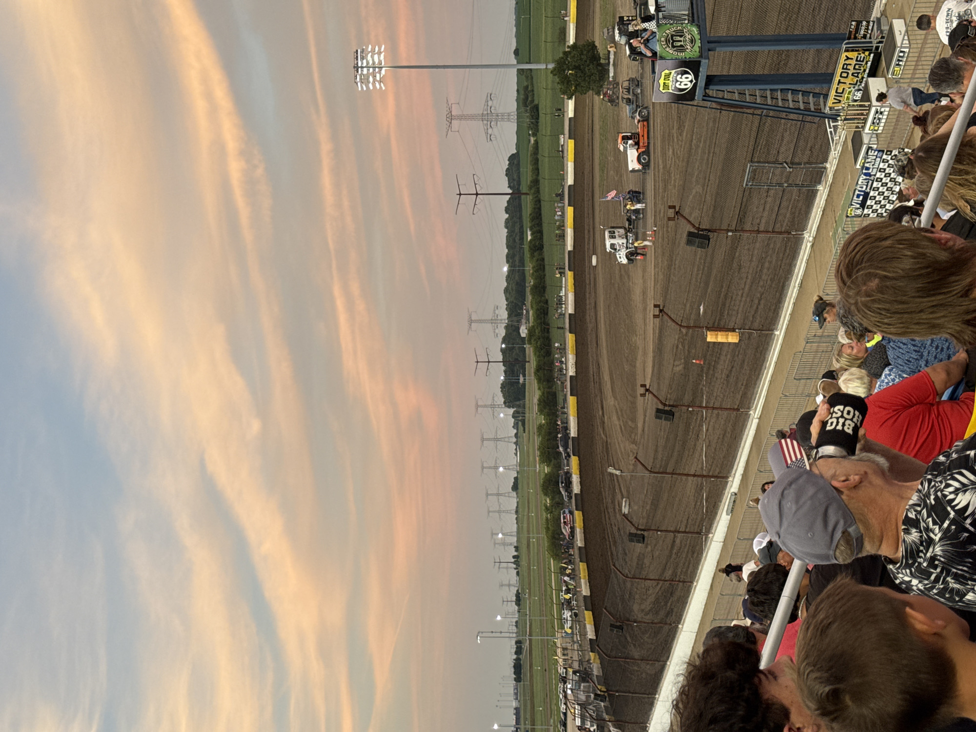 This is a medium-angle shot of the Dirt Oval at Route 66 Raceway in Joliet, Illinois, during a sunset or dusk event. The sky displays soft, peach and blue hues from the setting sun, with wispy clouds. In the foreground, a crowd of spectators are seated in bleachers, some holding drinks and wearing baseball caps. A man in a black and white patterned shirt and a gray cap with an American flag on it is visible in the mid-ground, holding a black can.
The race track, a dirt oval, occupies the central and lower portions of the image. Several vehicles, including trucks and what appears to be a tow truck, are positioned on the track and in the pit area. A tall light tower with bright stadium lights is illuminated on the right side of the track. In the background, a line of power transmission towers stretches across a flat, green landscape. The Victory Lane structure, adorned with trophies and signage for "Dirt Oval Route 66" and "Monster Truck," is prominent on the right side of the image, near the finish line. The overall scene depicts a racing event in progress or concluding, with a lively atmosphere among the spectators.