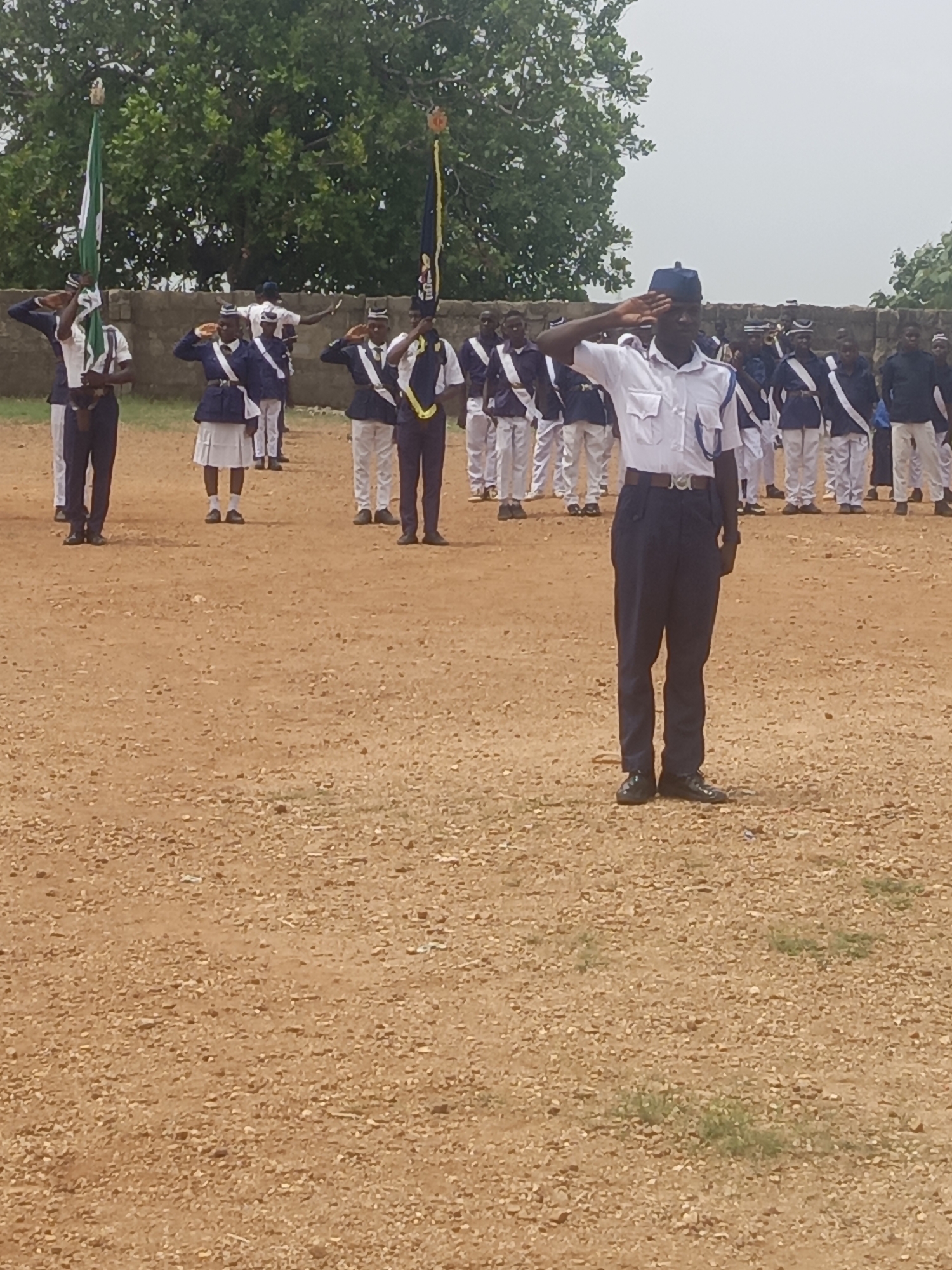 Akwanga, Nigeria: A group of approximately 20 individuals in what appear to be cadet uniforms are standing in formation on a brown, gravel-covered field. They are saluting. The uniforms are a mix of dark blue jackets and trousers, white shirts, and white skirts. Some have white and blue sashes. Two individuals are holding flags. In the background are green trees and a light-colored sky.