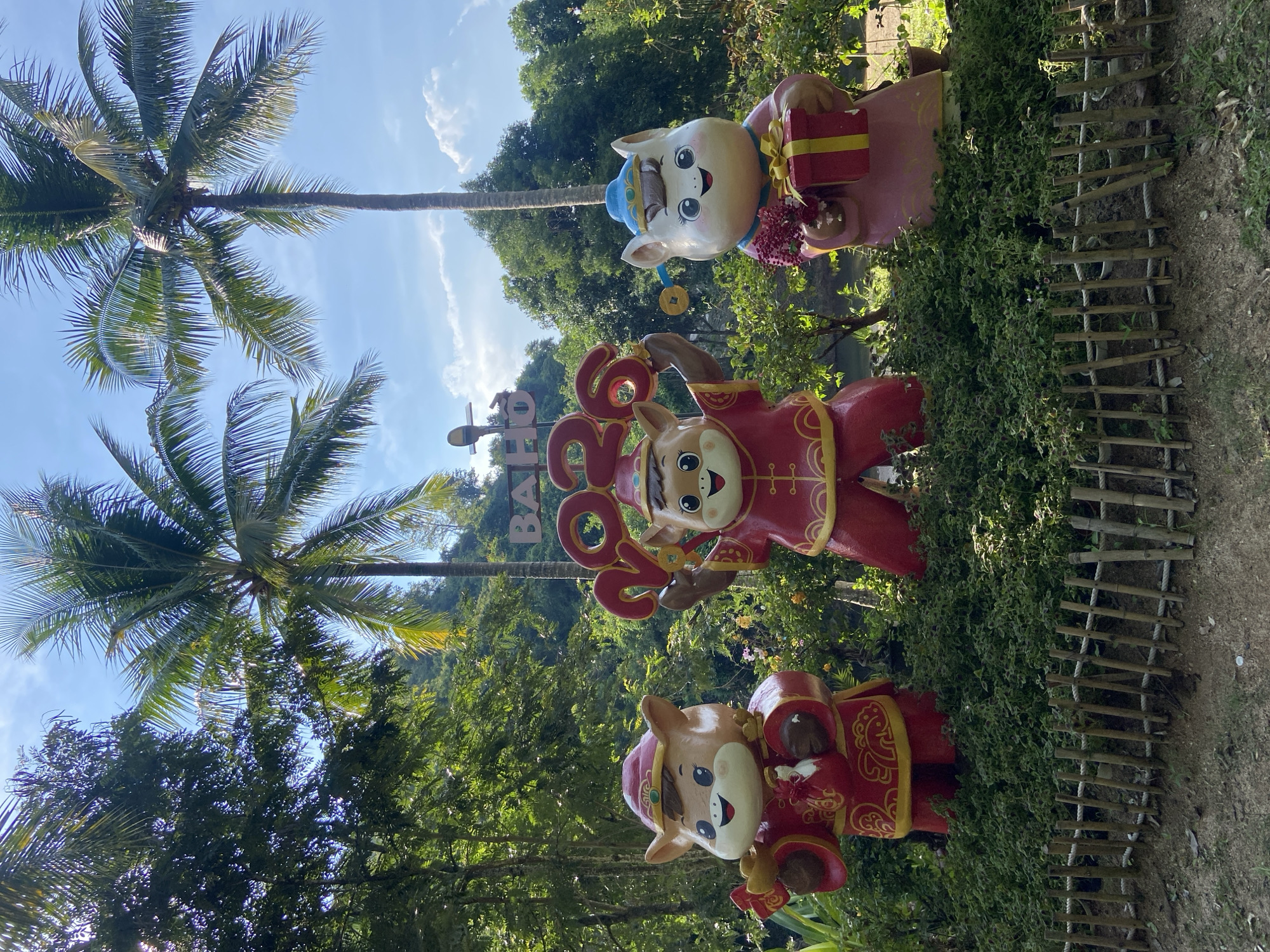 This is a medium shot of a festive outdoor display. Three cartoonish animal statues are arranged in a row, adorned in traditional red and gold Chinese New Year attire. The statue on the left, a brown bovine with a red hat, holds a golden coin and a small red pouch. The central statue, also a brown bovine, wears a red outfit and holds the digits "2023" above its head, signifying the year. The statue on the right, a light pink feline with a blue hat, clutches a bouquet of red flowers and a gift-wrapped box. They are positioned in front of lush green bushes and a wooden fence made of bamboo. In the background, palm trees and dense foliage are visible under a clear blue sky with scattered clouds. A sign with the word "BAHO" is partially visible behind the statues. The overall scene suggests a celebration or decoration in Hoa Thang Ward, Vietnam.