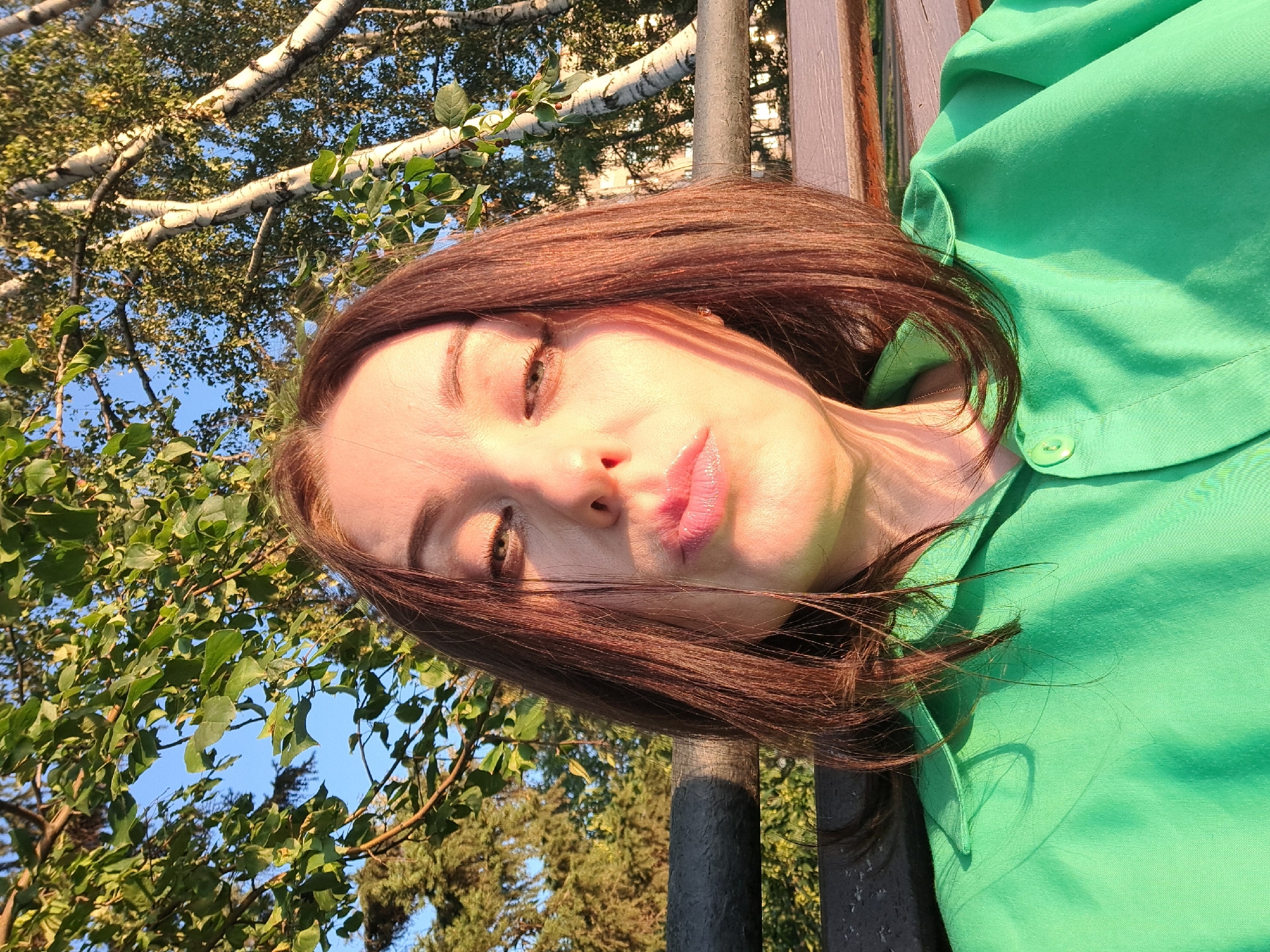 A woman with shoulder-length brown hair is taking a selfie outdoors. She is wearing a bright green collared shirt and is sitting on a wooden bench. The background shows trees with green leaves and white bark, and a clear blue sky. The sun is casting shadows on her face and shirt. There is no identifiable information to determine the location in the city of Irkutsk, Russia.