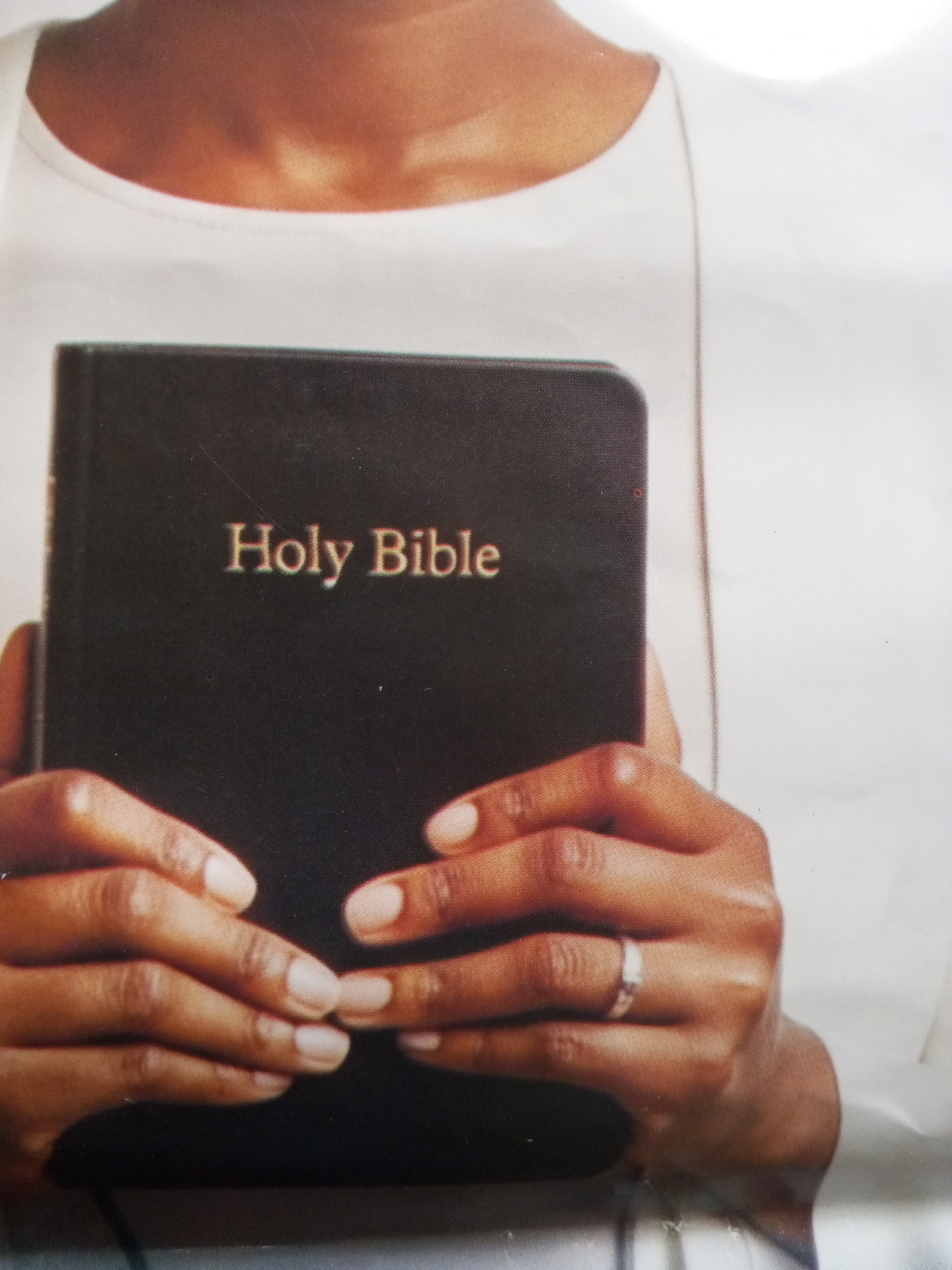 A close-up image captures a person, likely in Badagry, Nigeria, holding a black "Holy Bible." The individual has brown skin and is wearing a white garment, visible around the neck and shoulders. Both hands are positioned centrally, firmly grasping the Bible. The book's cover is dark, featuring the title "Holy Bible" prominently displayed in gold or bronze capital letters. A simple, light-colored band ring adorns the fourth finger of the person's right hand, and the fingernails are neatly manicured with a light, neutral polish. The background is a plain, indistinct white, suggesting an indoor or studio setting. The photograph exhibits a visible dot matrix pattern, indicating it may be a photo of a printed image, and minor dust specks or surface imperfections are present on the book and background. The focus is sharp on the hands and the Bible, highlighting the central object and the act of holding it.