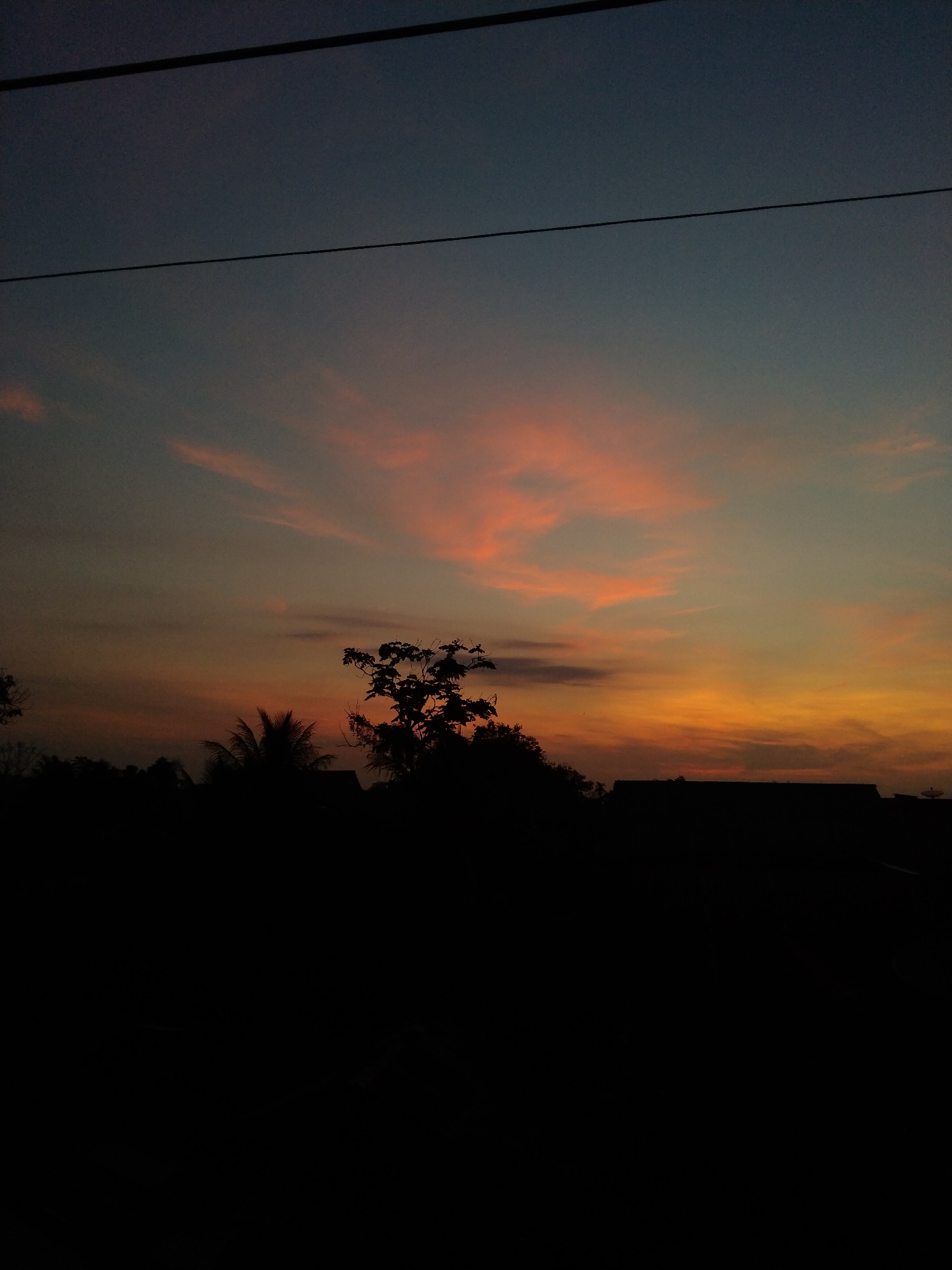 A low-angle, long-shot photograph captures a twilight scene. Overhead, two thin black electrical wires stretch horizontally across the top portion of the frame. The sky transitions from a dark blue in the upper region to orange and pink hues near the horizon. Patches of reddish-orange clouds are visible. The silhouetted outlines of trees and buildings form a dark horizon line. The photo was taken in Bireuenmeunasahdayah, Indonesia.