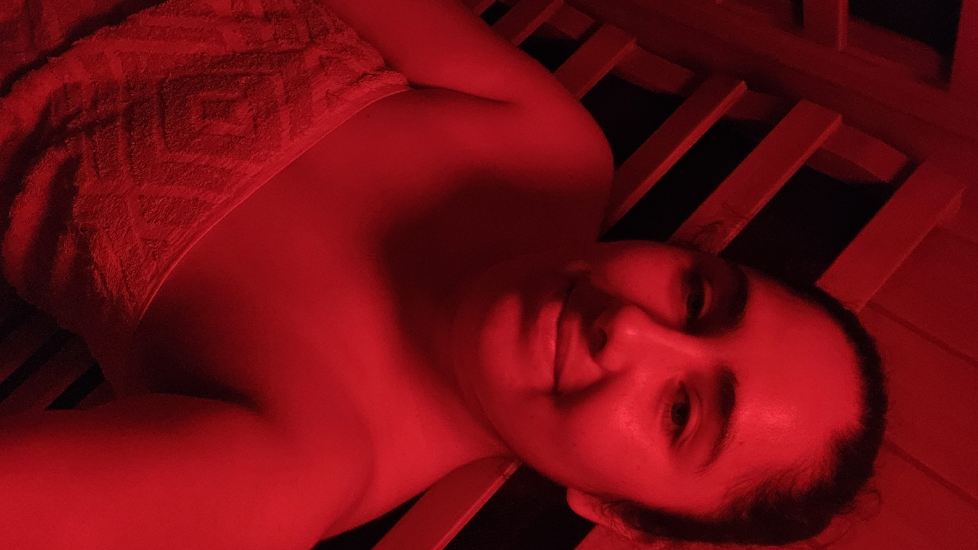 The media file presents a portrait of an individual lying in an environment saturated with intense red light. The subject, an adult, is positioned diagonally across the frame, with their head and upper torso visible. A red, patterned towel or blanket is draped over their body, covering the chest and shoulders. The person has a subtle smile on their face, indicating a sense of relaxation or contentment.
The setting appears to be an indoor space, likely an infrared sauna or a red light therapy bed, given the uniform red illumination and the visible wooden slats that form a structure around and beneath the person. The red light casts deep shadows and highlights contours, creating a striking monochrome effect where all visible elements are variations of red, black, and deep red tones. The background behind the slats is dark, suggesting an enclosed space. The overall activity seems to be a session of relaxation or therapeutic treatment. No visible text is present in the image. The location context provided is Puebla, Mexico.