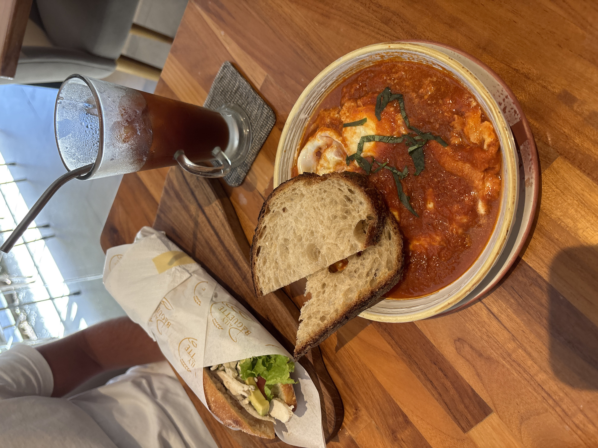 The image depicts a meal arrangement on a wooden table, featuring a beverage, a sandwich, and a dish of shakshuka with bread. A tall glass of iced tea with a straw sits on a gray coaster to the left. Next to it is a sandwich wrapped in branded paper labeled "The Daily Baguette," revealing ingredients such as lettuce, avocado, and chicken. Adjacent to the sandwich, two slices of bread accompany a bowl of shakshuka topped with green herbs, likely basil. The setting appears to be a cafe or restaurant in Denpasar, Indonesia, implied by the dishes served and the style of the establishment. An individual wearing a white shirt is partially visible in the lower left corner.