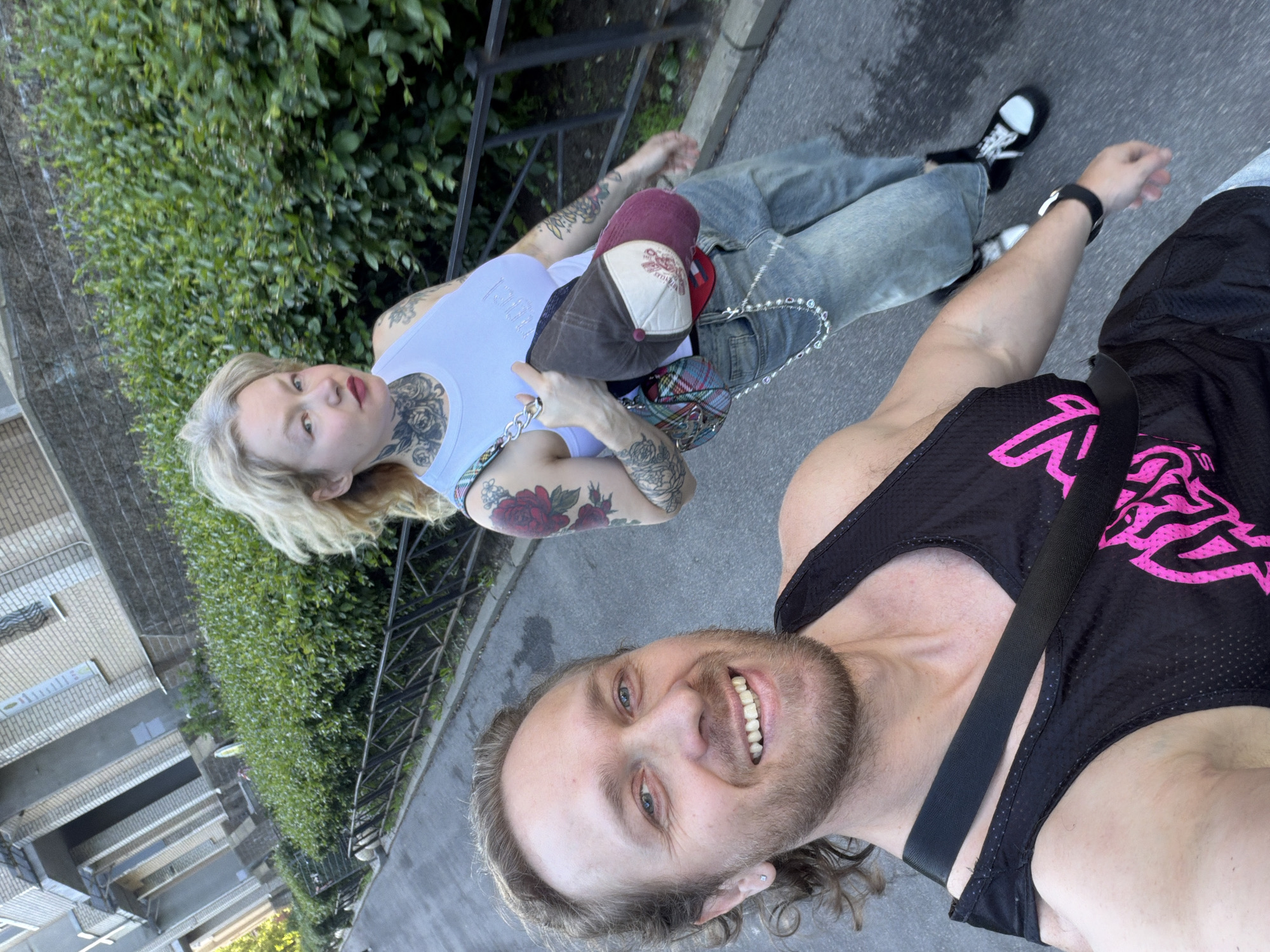 An outdoor, upward-angled selfie-style photograph captures two individuals walking on a paved surface in Saint Petersburg, Russia, likely a sidewalk adjacent to a road. The scene is illuminated by natural daylight.
In the foreground, slightly to the left, a light-skinned male with medium-length curly brown hair and a short beard smiles directly at the camera, displaying his teeth. He has light blue eyes and wears a black sleeveless athletic tank top with a partial pink graphic design. A black strap, likely from a bag, crosses his right shoulder. A small silver square stud earring is visible in his left earlobe, and a black watch is on his left wrist.
Positioned to the right and slightly behind the male, a light-skinned female walks with her gaze directed generally forward and slightly left. She has windswept blonde hair and prominent tattoos, including a floral design on her neck, a large red rose and other patterns on her left arm, and multiple designs on her right arm. She wears dark red lipstick. Her attire consists of a white sleeveless tank top and blue-wash wide-leg jeans with a silver chain accessory hanging from the right side. A small, multi-colored plaid handbag with a silver chain strap is slung over her left shoulder. She holds a two-tone baseball cap (brown on top, white and maroon on the front) in her left hand, tucked under her arm.
The background features a low, black metal fence running parallel to the path, behind which is a dense, well-maintained green hedge. Above the hedge, light-colored, multi-story brick buildings with multiple windows and distinct architectural details, consistent with residential structures in an urban environment, are visible. One building exhibits a sign containing Cyrillic script. The paved surface beneath the individuals is dark grey.