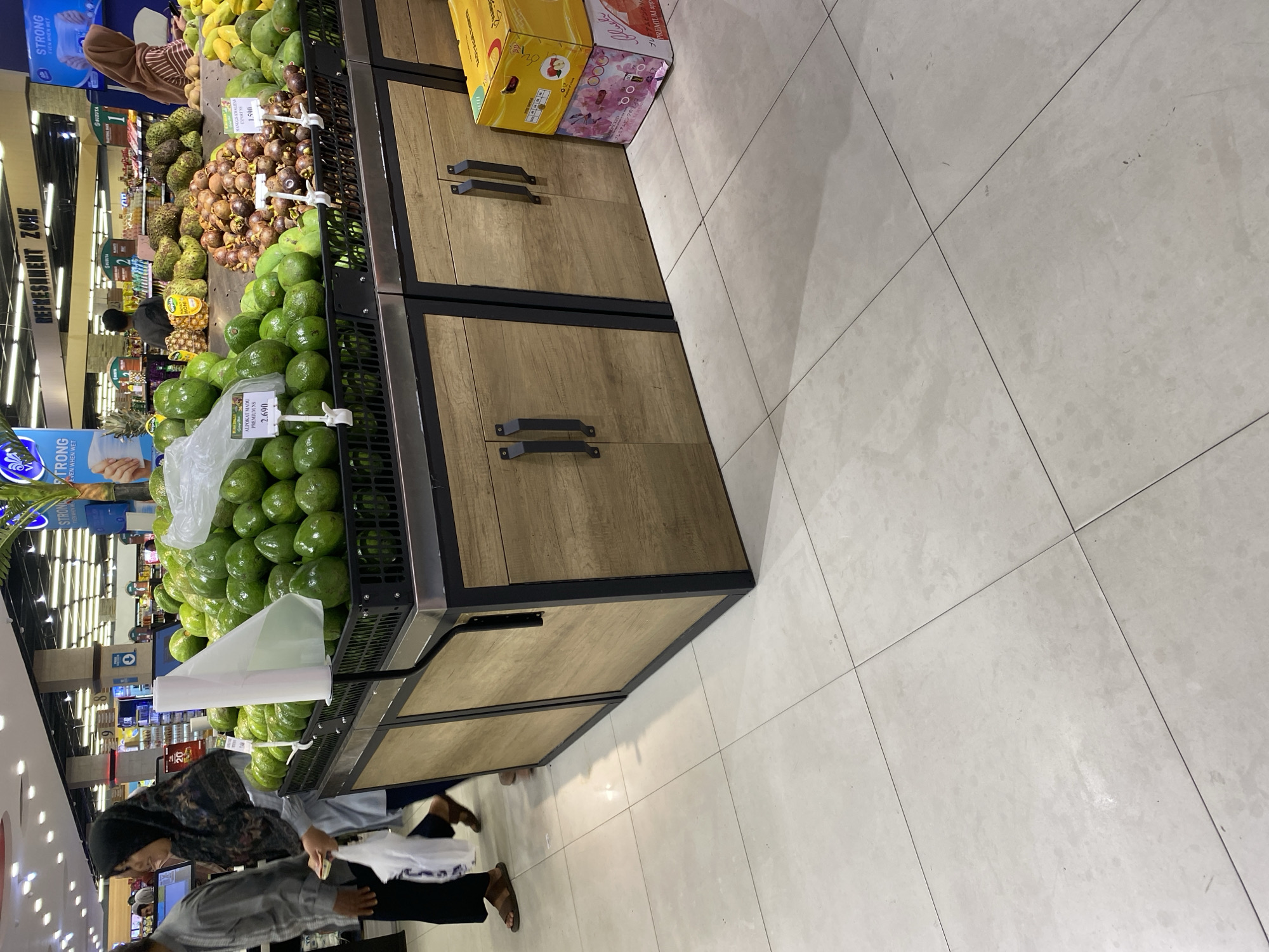In the produce section of a supermarket in Banda Aceh, Indonesia, a display of ripe avocados is prominently featured in black plastic crates. Behind the avocados, a variety of other fruits are visible, including soursop, mangosteen, and mangoes. Two individuals are seen walking past the fruit display, one a woman in a headscarf and the other a man. The supermarket's interior, with its tiled flooring and overhead lighting, is also visible. The overall scene depicts a typical shopping environment in a tropical region.