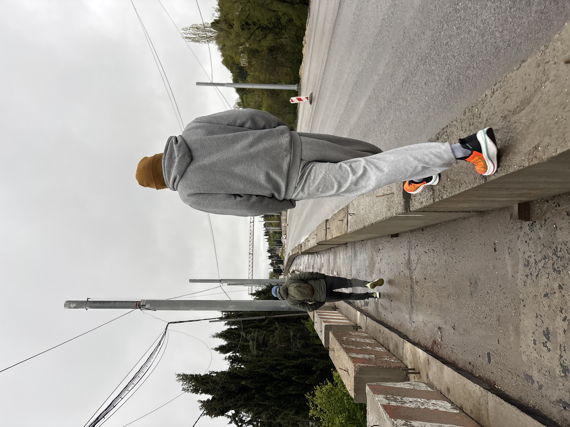 The image depicts an elevated concrete walkway or bridge in Kiparisnoe, Ukraine, under an overcast sky. Two individuals are visible. One individual, closer to the viewer, is dressed in a gray sweatsuit and a brown beanie, with orange and black athletic shoes. This person is standing on the edge of the walkway. Further in the distance, another person is walking along the walkway. They are wearing a jacket, carrying a backpack, and have on a blue head covering. Utility poles line the walkway, with wires extending across the scene. The walkway appears to be bordered by trees and vegetation. The road surface and concrete appear wet.