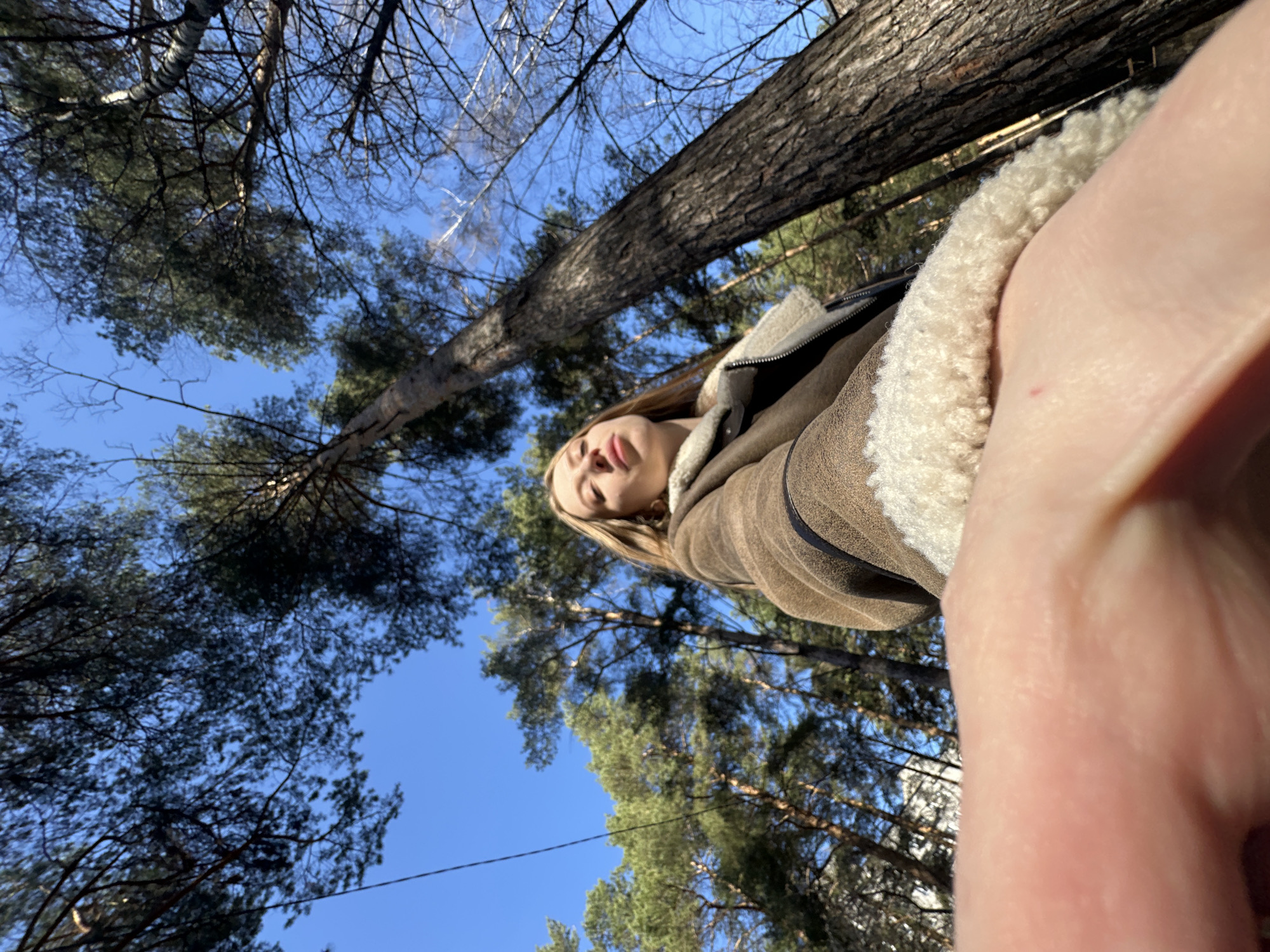 The image is taken from a low-angle perspective, looking up towards a person wearing a brown coat with a white, fluffy lining. The person's face is visible, and they appear to be looking towards the camera. The background consists of trees and a blue sky. The location is likely a wooded area. Given the prompt specifies Chaikovsky, Russia, the wooded area is assumed to be within the city limits or its immediate surrounding areas.