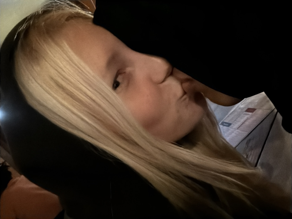 A blonde woman with fair skin pouts her lips toward an unseen person or object. She wears a dark hooded sweatshirt. Her long, straight hair is parted to one side and falls across her face. The lighting is dim, creating shadows on her face and hair. A partial view of a patterned document or surface is seen near the unseen person or object she is pouting toward.
