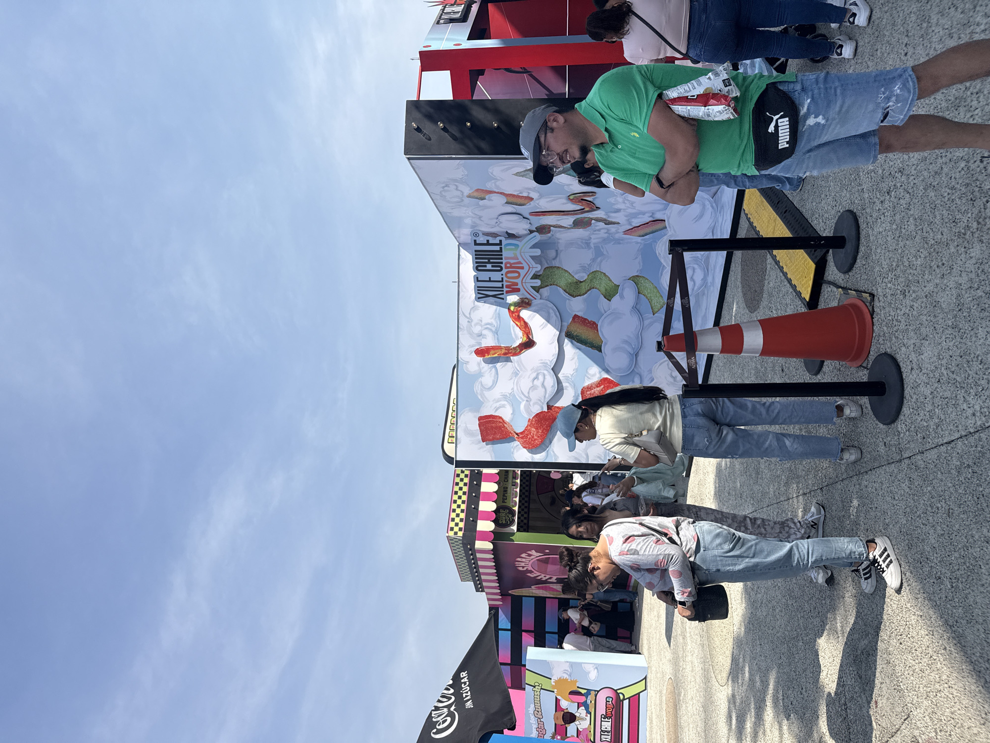 The image shows a group of people standing in line at an event called "Xile Chile World", as indicated by the large signage behind them. The sky is blue and relatively clear. The people are of various ages and genders. One man is wearing a green polo shirt and denim shorts with a black Puma fanny pack. He is also wearing a grey baseball cap and glasses and appears to be looking at something. Another person is wearing a light-colored shirt and jeans. The third person in line has on a patterned shirt and jeans. An orange traffic cone is placed behind them. The event appears to have booths decorated with colorful designs, including a Coca-Cola banner and a section labeled "Sugar Ruuush". The location is likely Mexico City, Mexico, considering the name and signage written in Spanish.