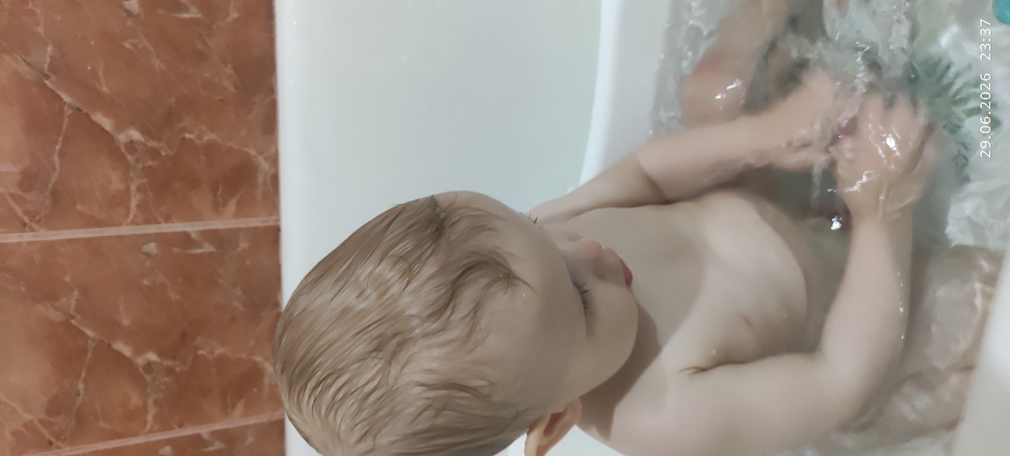 An overhead shot shows a young child, likely a toddler, bathing in a tub. The child is fair-skinned with light brown, wet hair. They are looking down at their hands, which are submerged in water, creating ripples. The child's body is mostly submerged in the water, with only their upper torso and head visible. The background consists of tiled walls and the white edge of the bathtub. The timestamp "29.06.2026 23:37" is visible in the bottom right corner of the image.