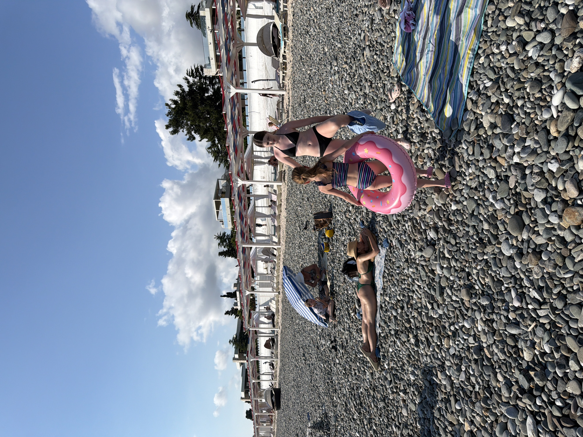 The image depicts a crowded pebble beach under a bright, partly cloudy sky. Several people are seen enjoying a sunny day at the beach. In the foreground, two young girls are playing, one sitting and holding a pink inflatable ring, and the other standing. Behind them, an adult woman in a black bikini is partially visible. Further back, several other individuals are scattered across the beach, some lying on towels or sunbathing. There are beach umbrellas and lounge chairs arranged in rows along the shore, suggesting a developed resort area. The sea is not directly visible, but the beach environment implies it is nearby. The overall mood is relaxed and leisurely, characteristic of a summer day at the beach. The time of day appears to be midday or early afternoon, given the bright sunlight.