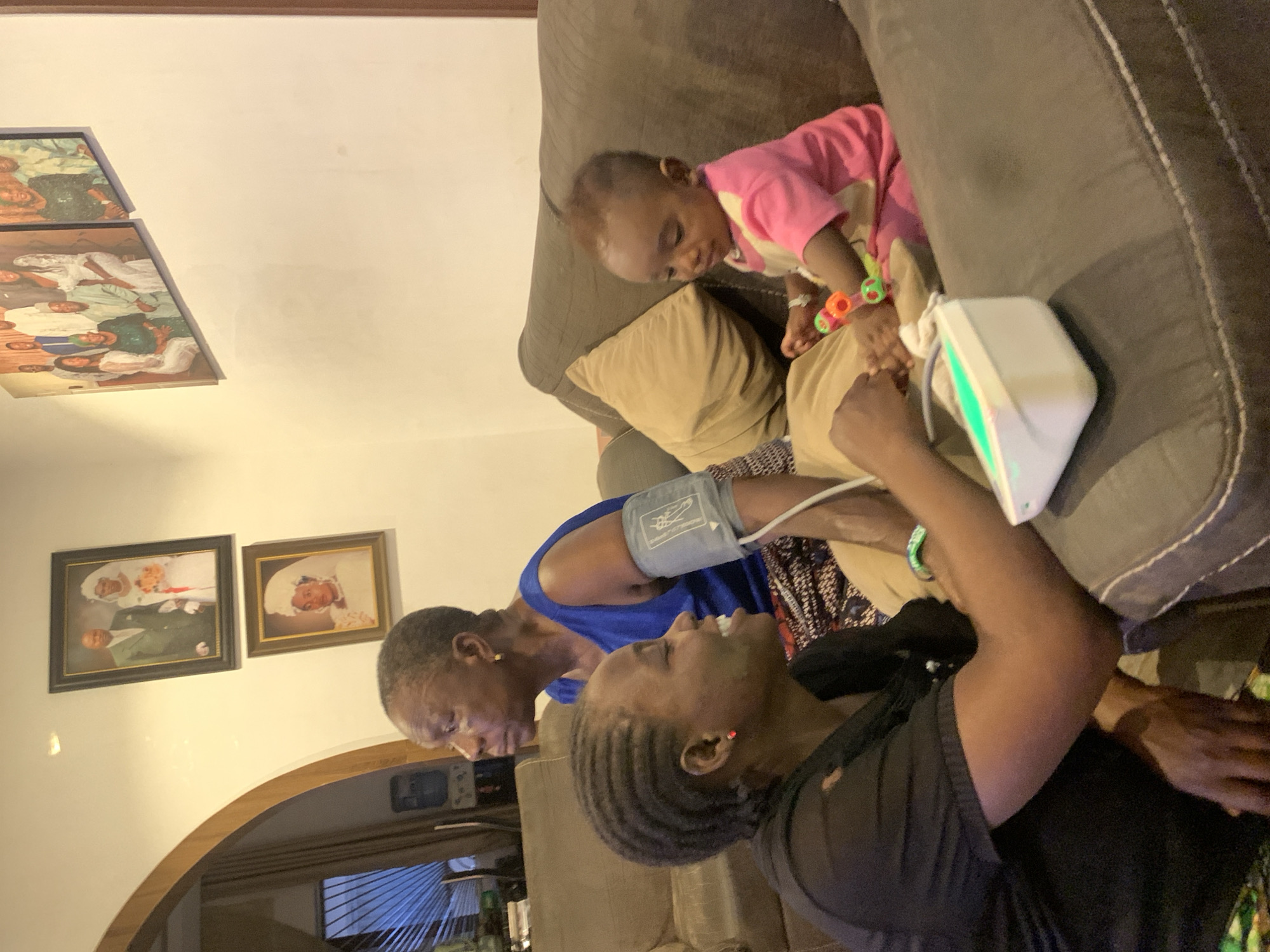 A family is situated on a brown sectional sofa in an indoor environment. An infant in pink and white clothing sits on the sofa with a colorful toy around their ankle. A woman in a blue top has a blood pressure cuff on her arm, while another woman in a black top operates a blood pressure monitor. The location is identifiable as Jos, Nigeria, due to the distinctive interior design and cultural elements. Paintings on the wall, including what appears to be a wedding portrait, add to the domestic atmosphere.