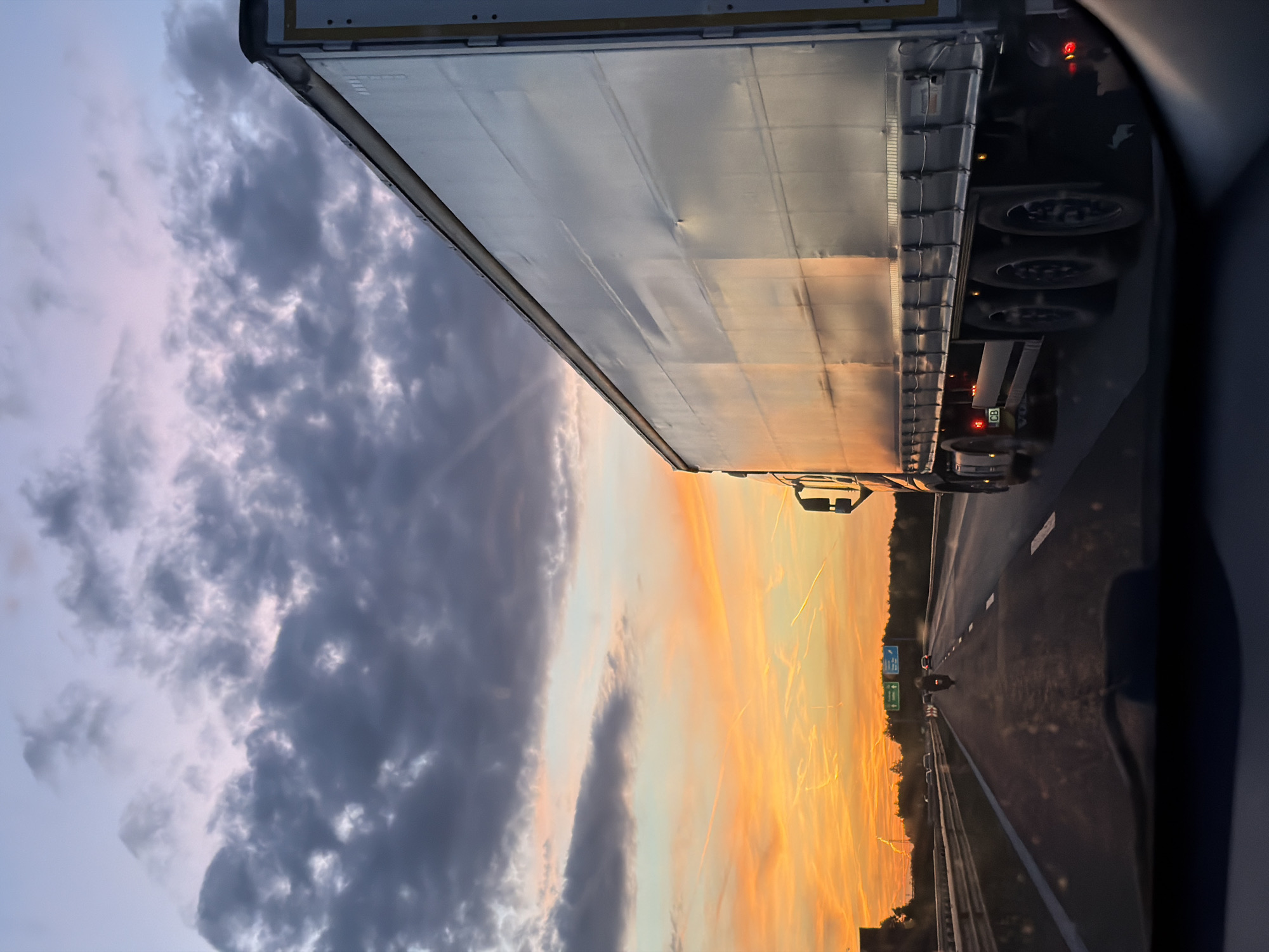 The image depicts a view from a vehicle moving on a highway. A large truck is positioned to the right of the viewer's vehicle. The background features a vivid sunset with orange and yellow hues blending into a blue and cloudy sky. The road shows other vehicles ahead. The location is identified as Baliovtsi, Bulgaria. The scene suggests ongoing travel during twilight.